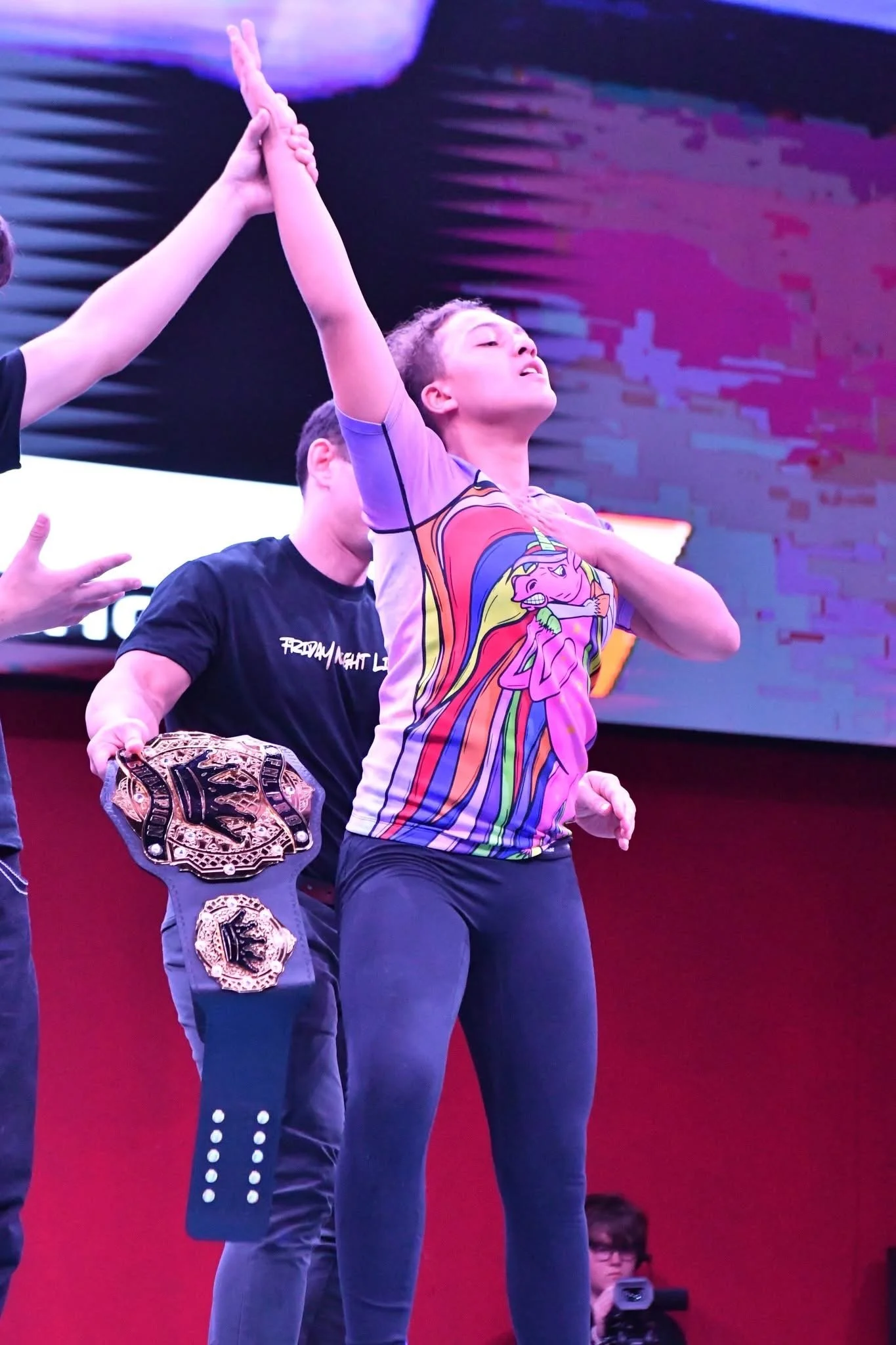 A female athlete with her eyes closed, one hand on her chest, wearing a colorful 'My Little Pony' shirt, standing confidently with her arm raised and clasped with another person's hand during a ceremony. The person next to her holds a championship belt.
