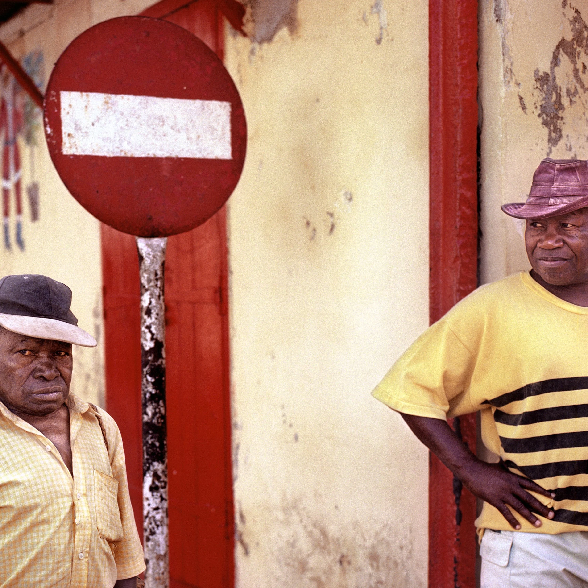 In Rodrigues Island, POrt Mathurin, Two men standing next to a worn wall and a weathered red and white 'no entry' traffic sign; one man wears a checkered shirt and a cap, the other is in a yellow striped shirt and a hat.