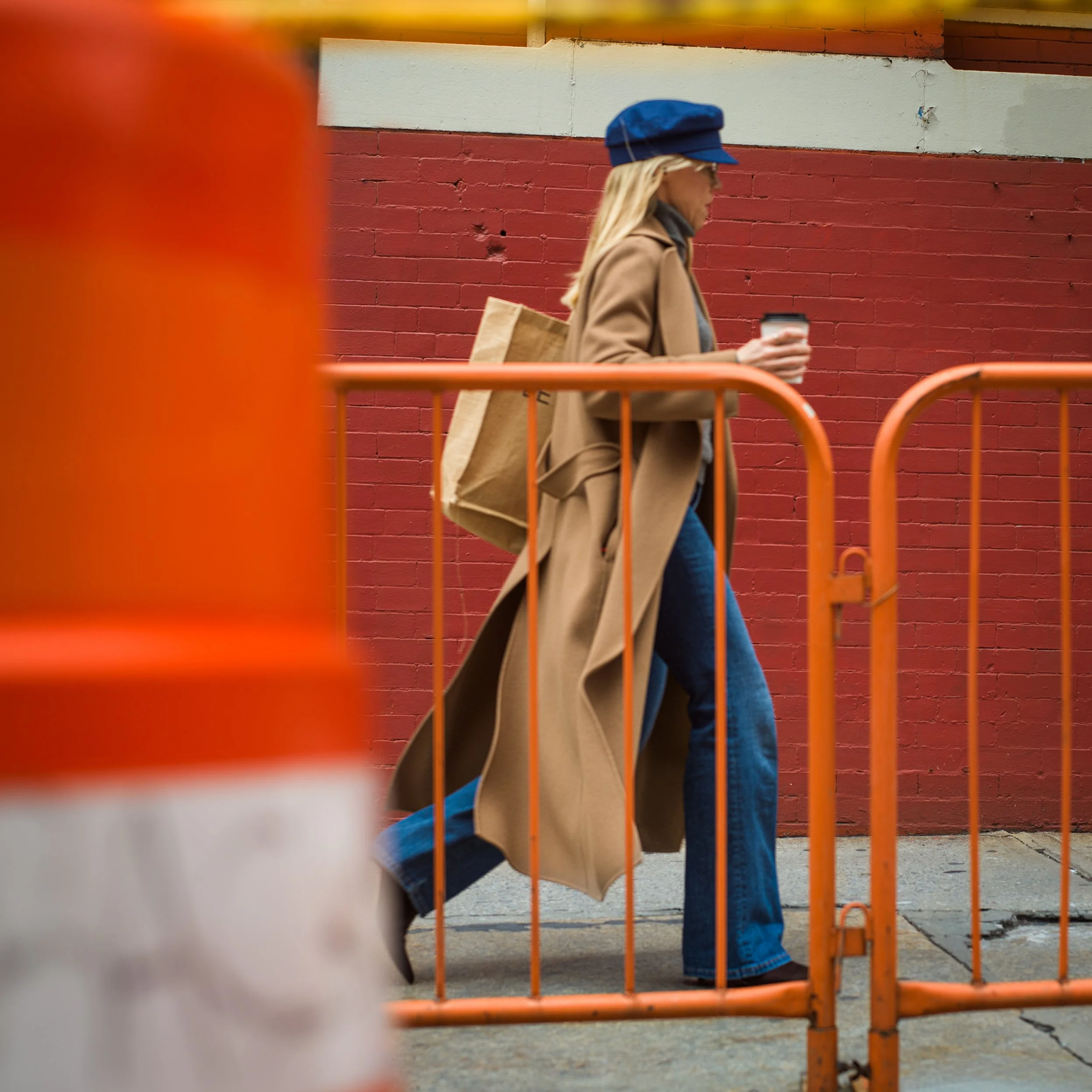 In New York Manhattan, A woman with blonde hair, wearing a blue cap, tan coat, and blue jeans, is walking along a sidewalk behind an orange safety barrier while holding a coffee cup. 