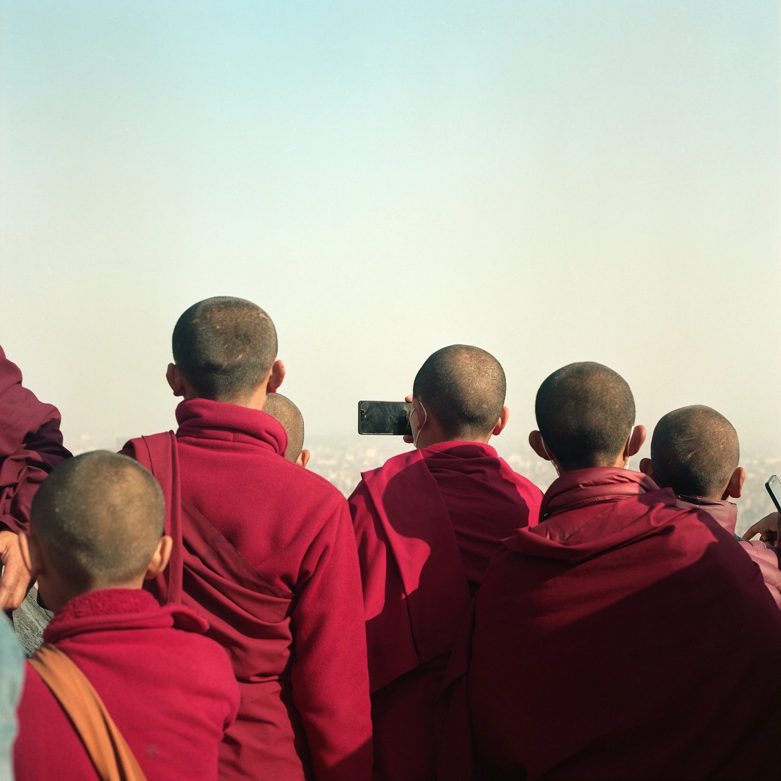 In Katmandu, A group of young monks in maroon robes, sitting together and taking photos with mobile phones, facing away from the camera into the distance.