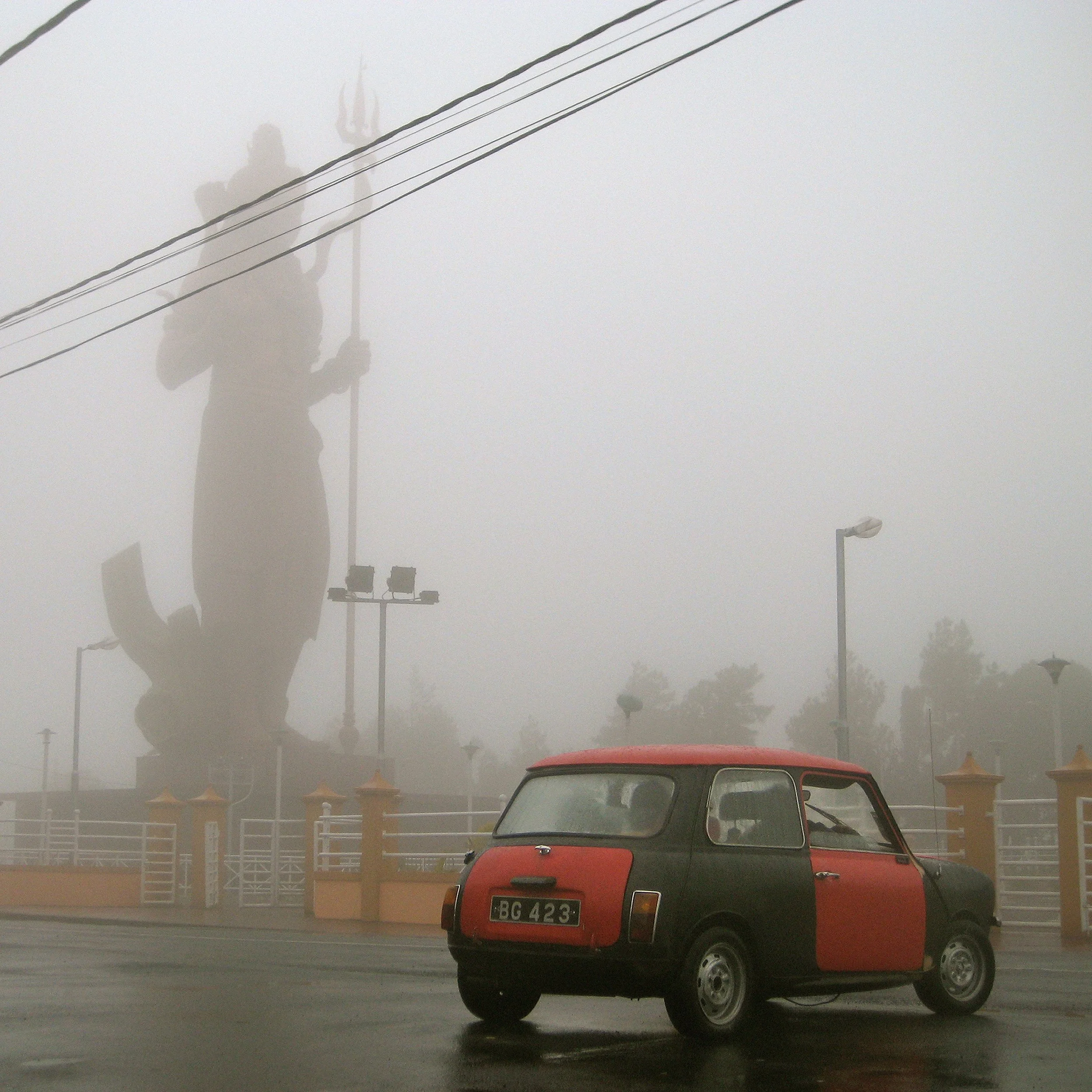 In Mauritius, Ganga Talao, An old mini cooper red classic car in a parking lot on a foggy day. In the background, a monumental statue of Shiva.