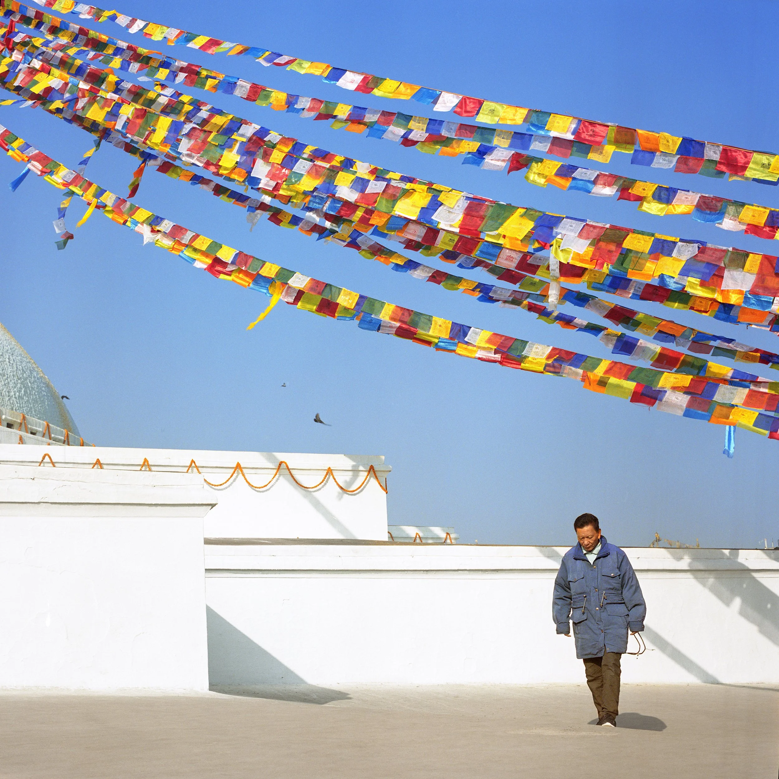 IN Katmandu, A man walking past white walls with prayer flags hanging above against a clear blue sky. The flags are colorful and flutter in the breeze.