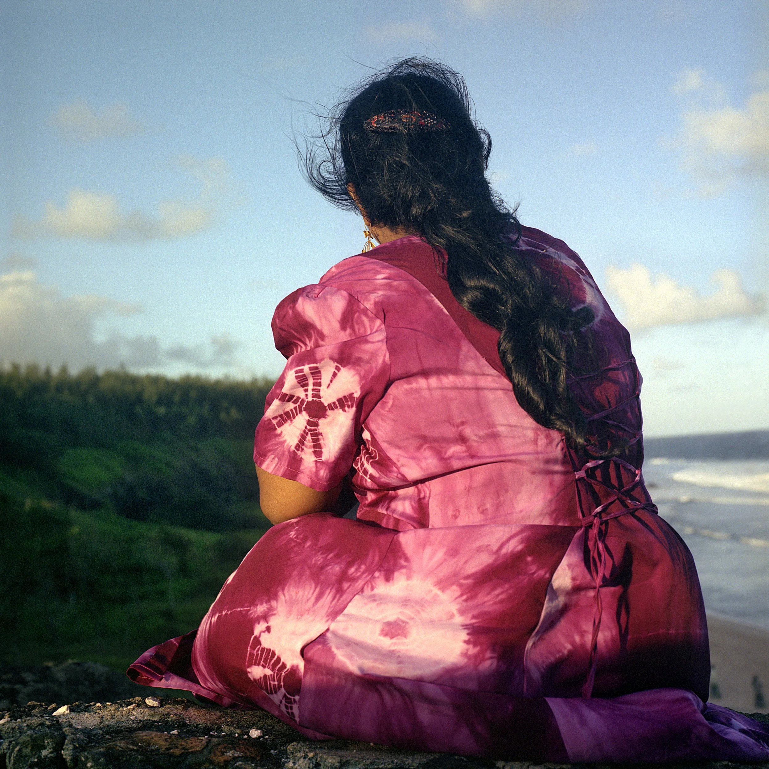 In Gris Gris Souillac Mauritius, A person with long, dark hair wearing a pink tie-dye shirt, sitting outdoors near a body of water and looking at the sky.
