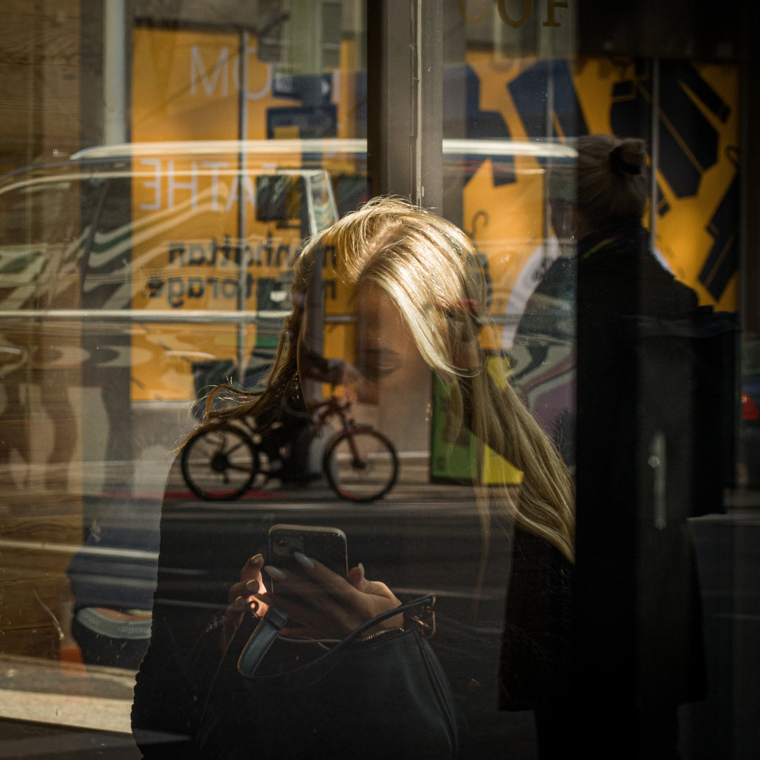 In New York Manhattan, Woman with blonde hair looking at her phone while sitting inside a coffee shop, reflected in the window with a bicycle icon and street scene reflection.