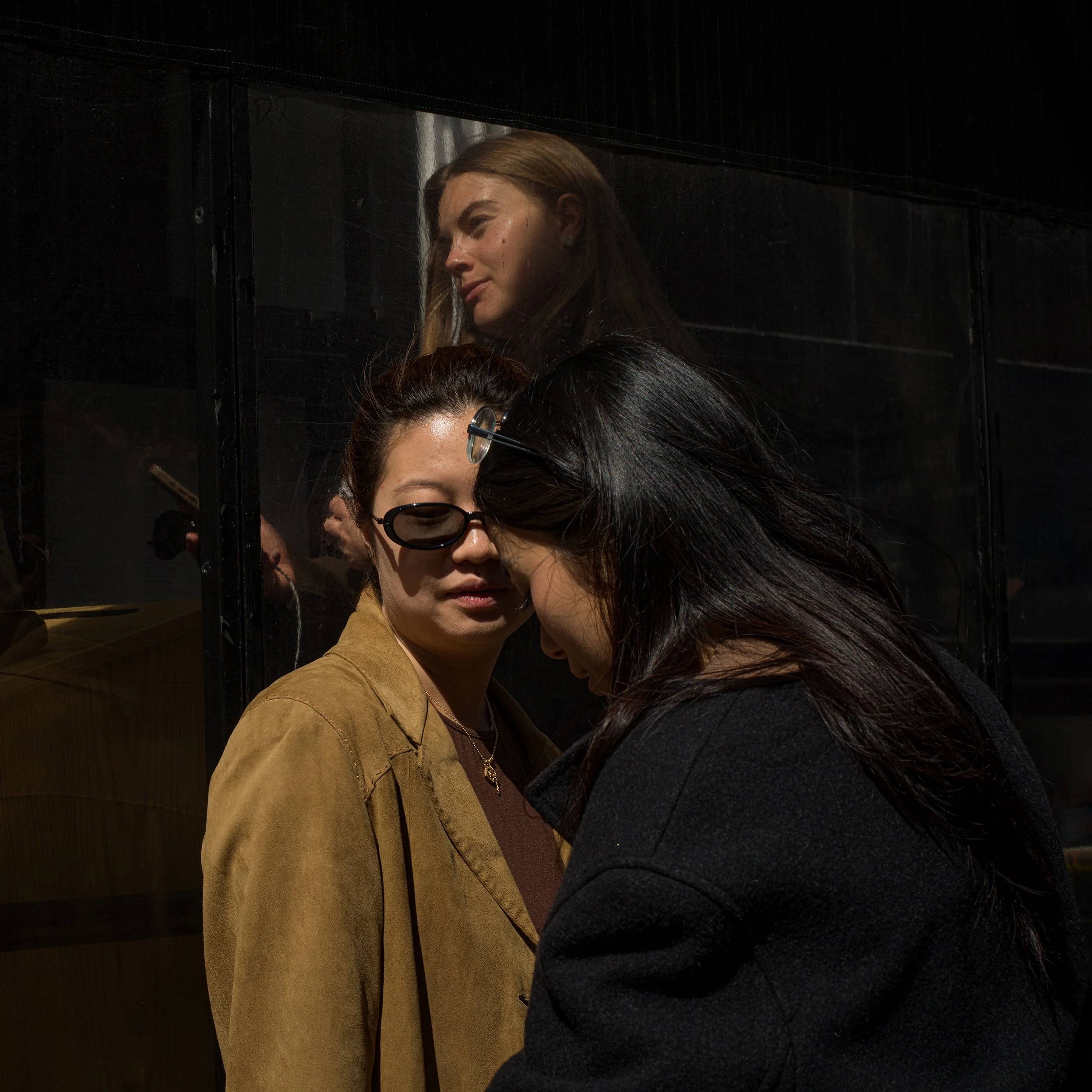 In New York Manhattan, Two women with dark hair, one wearing sunglasses and a brown jacket, are close to each other with foreheads touching. A third woman with blonde hair is reflected in a glass surface behind them, looking downward.