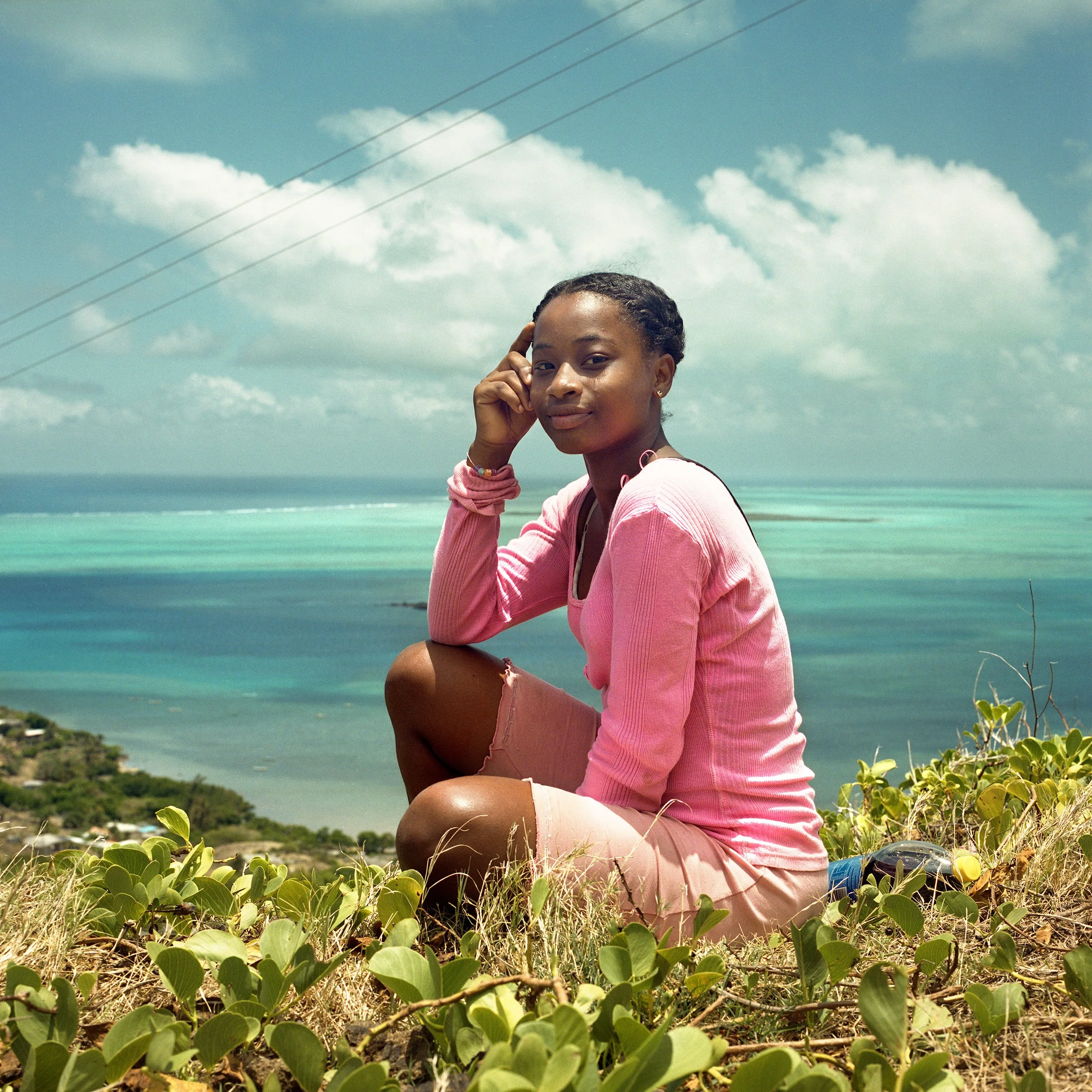 A woman sitting on a grassy hill overlooking the ocean, wearing a pink long sleeve shirt and pink shorts, with a calm expression.