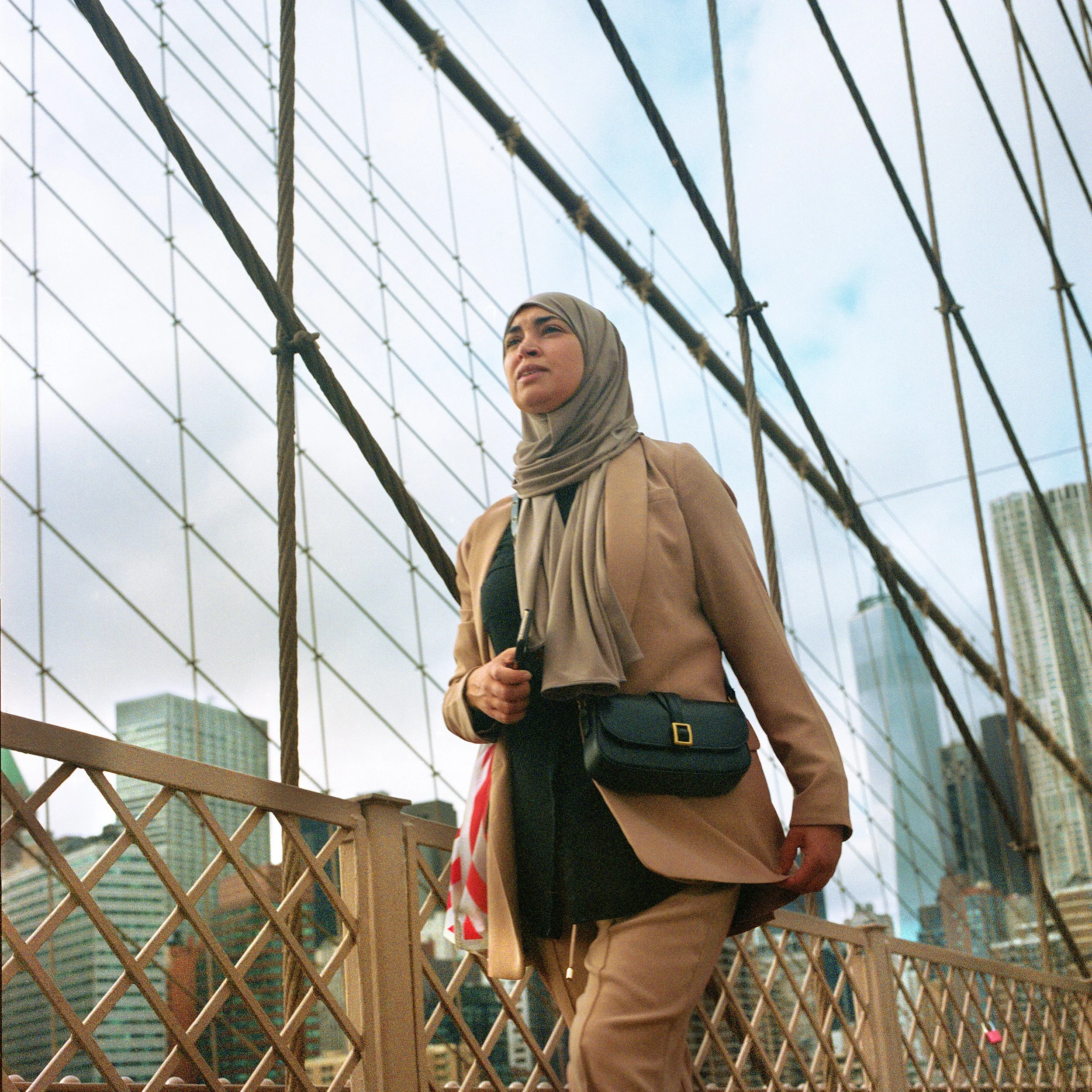 In New York Manhattan Brooklyn bridge, A woman wearing a beige hijab and coat standing on a bridge with city skyscrapers in the background, holding a camera and looking into the distance.