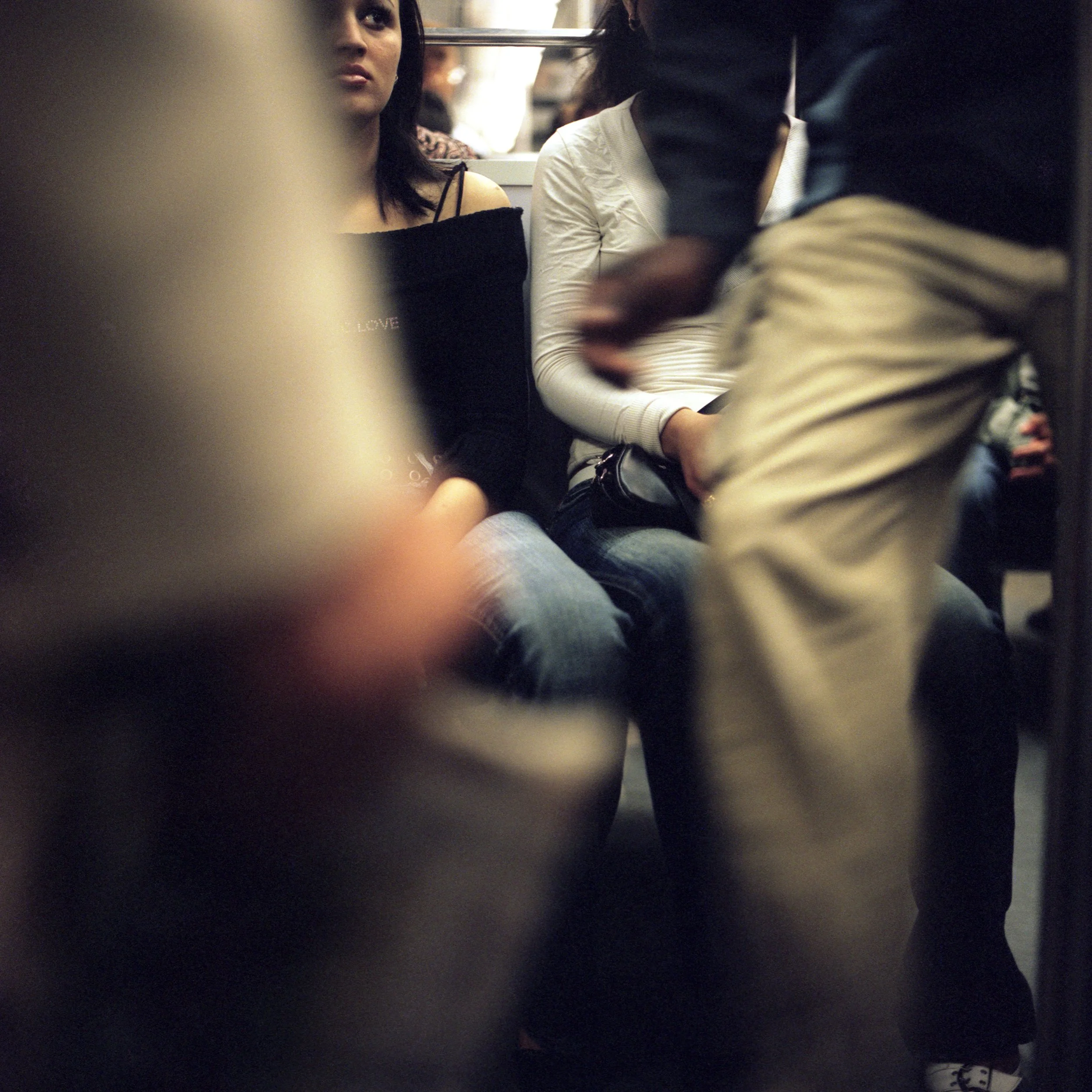 A woman with dark hair sitting in Paris Subway, looking straight ahead. She is wearing a black off-the-shoulder top and jeans. Another person is standing nearby, partially visible, wearing beige pants and a dark jacket.