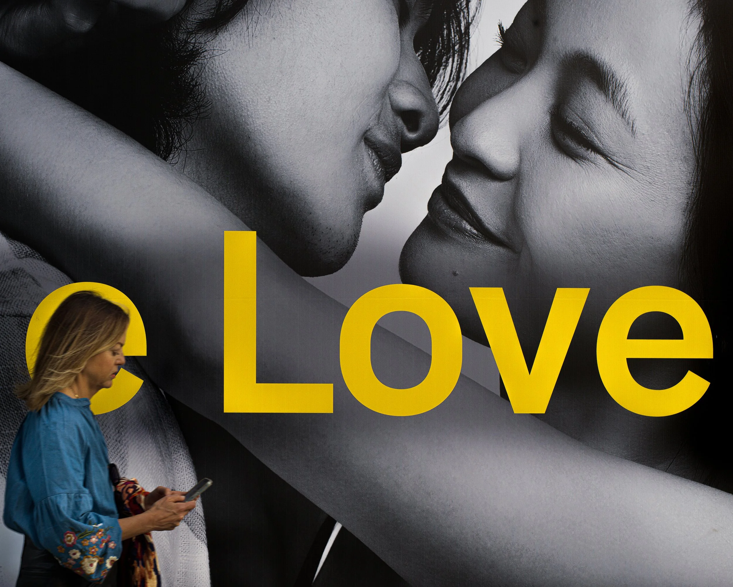 In New York Manhattan, A woman in a blue floral embroidered jacket looking at her phone, walking in front of a large black and white photographic advertisement of a couple embracing, with the word 'Love' in bold yellow letters across the image.