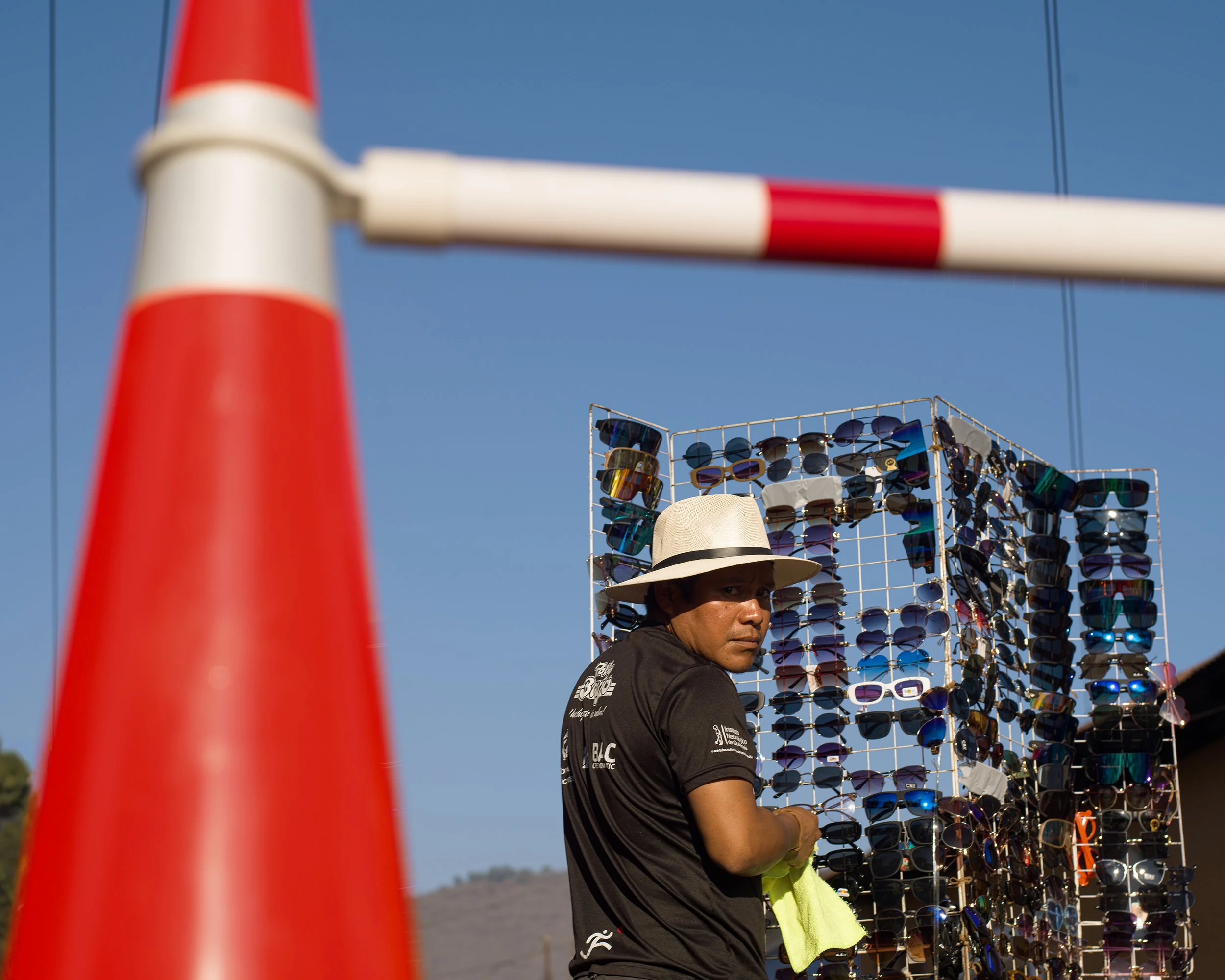 In Antigua Guatemala, A man wearing a wide-brimmed hat looks over his shoulder at a sunglasses stand, with a traffic cone in the foreground and a blue sky in the background.