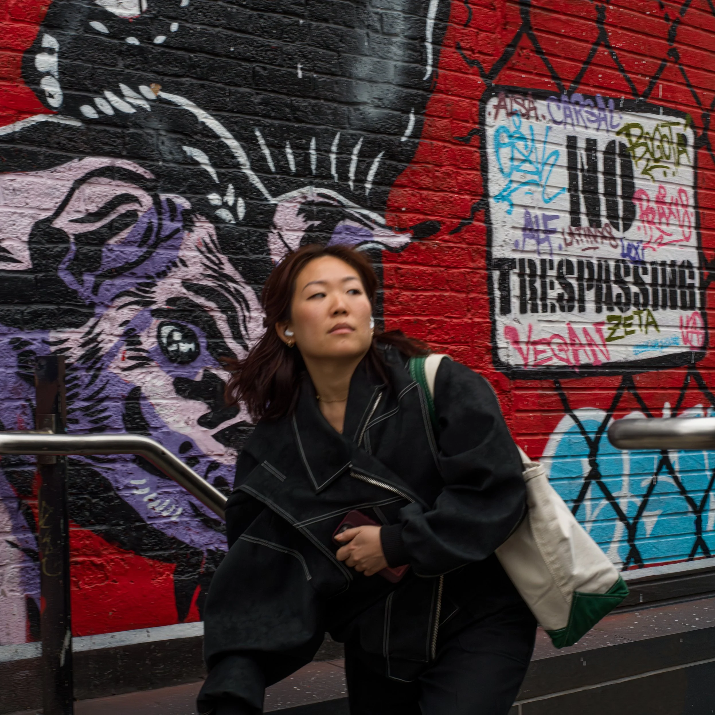 	
In New York Manhattan, A woman with brown hair wearing a black jacket and carrying a beige tote bag stands in front of a colorful mural on a brick wall. The mural features a tiger with graffiti tags.