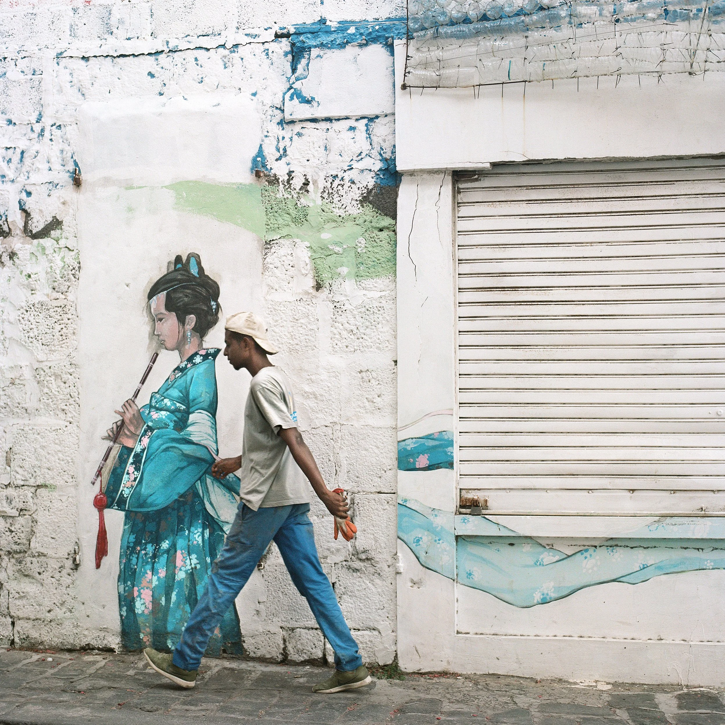 A man walking past a mural of a woman in traditional Asian attire, playing a flute, painted on a textured wall