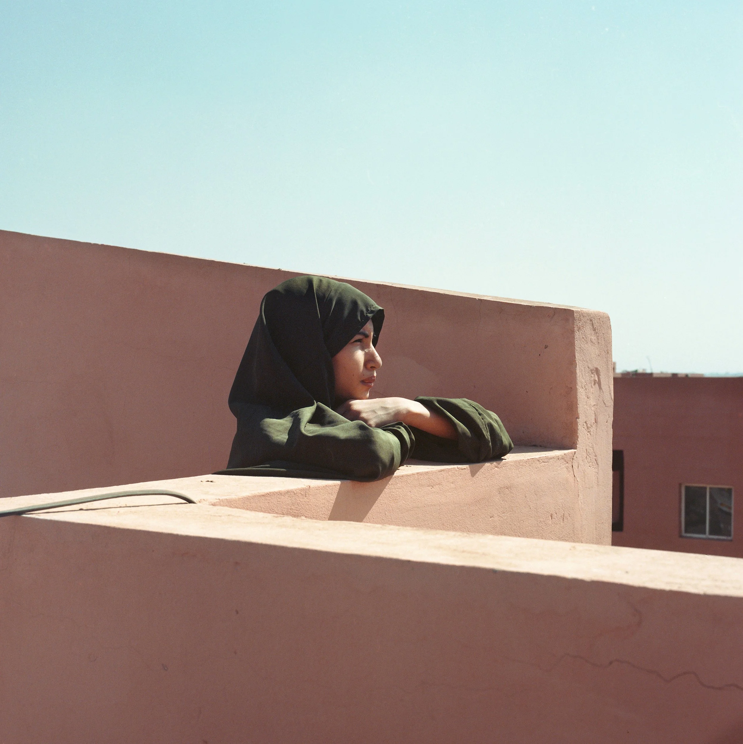 Young woman in a dark green hoodie resting her arms on a pink balcony wall and looking to the right under a clear blue sky.
