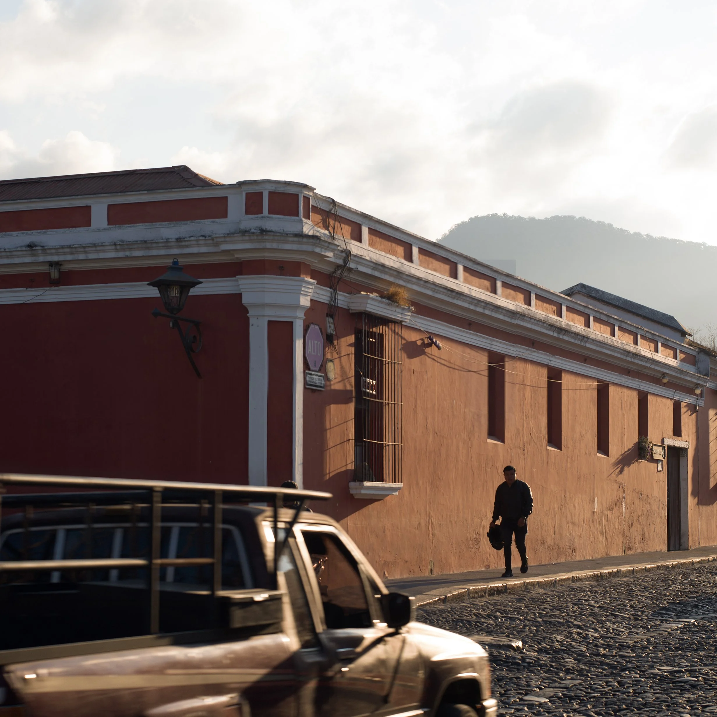 	
In Antigua, Guatemala, A person walking on a cobblestone street beside an orange building with barred windows, with a vintage black vehicle in the foreground and mountains in the background.