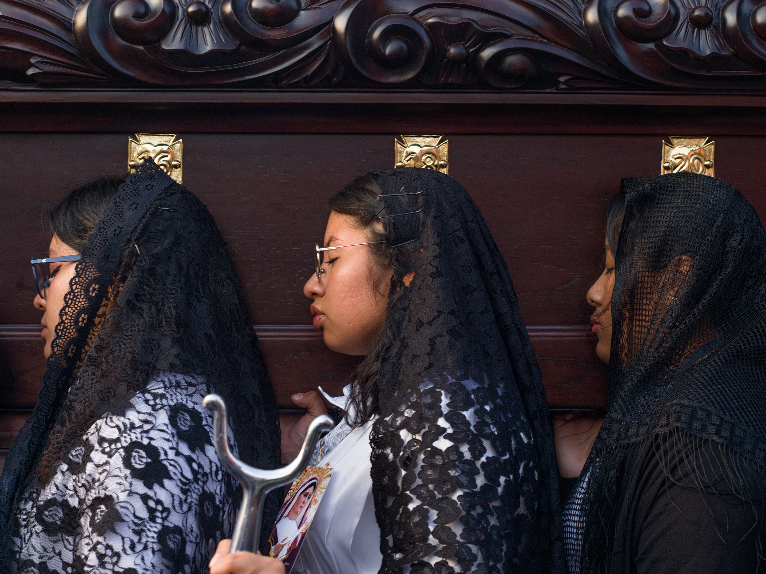 Antigua. Guatemala, Alfombras, Three women in black lace veils and glasses, with one holding a small image and a silver umbrella handle, in profile against a wooden background.