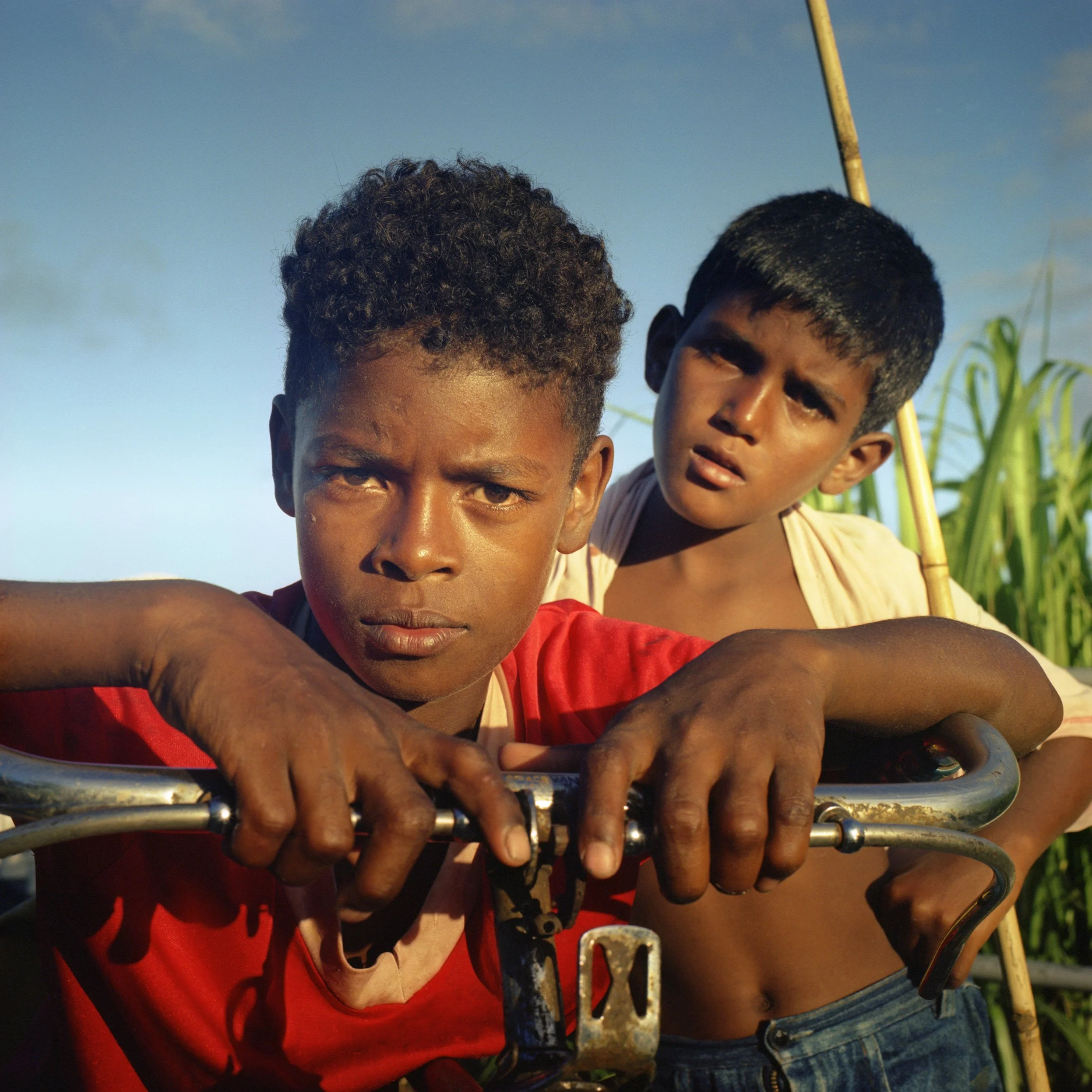 in Mauritius, Trou d'Eau Douce, Two young boys lean on a bicycle, looking directly at the camera with serious expressions, with green plants and a blue sky in the background.