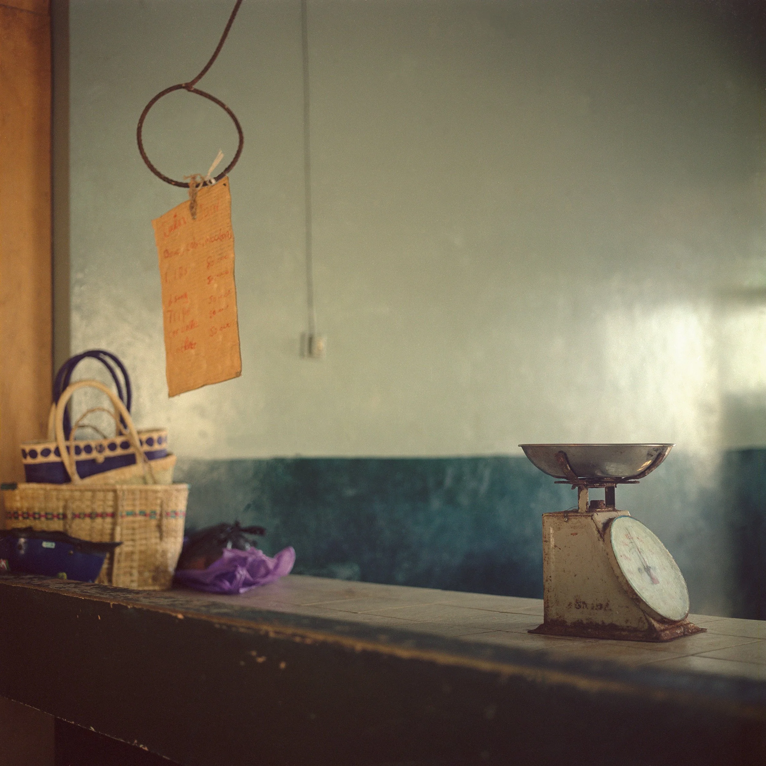 In Rodrigues Island, Mauritius, A kitchen scene with a vintage scale on a wooden counter and a hanging bundle of lip balm tubes in a basket, with bags and purple fabric at the bottom.