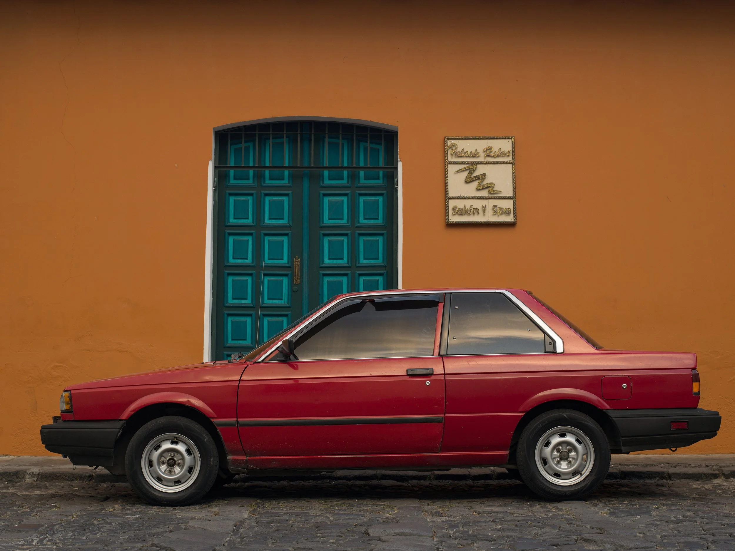 In Antigua, Guatemala, A red vintage two-door car parked in front of an orange building with a blue door and a sign that reads 'Palacio Rivas Salon y Spa'.