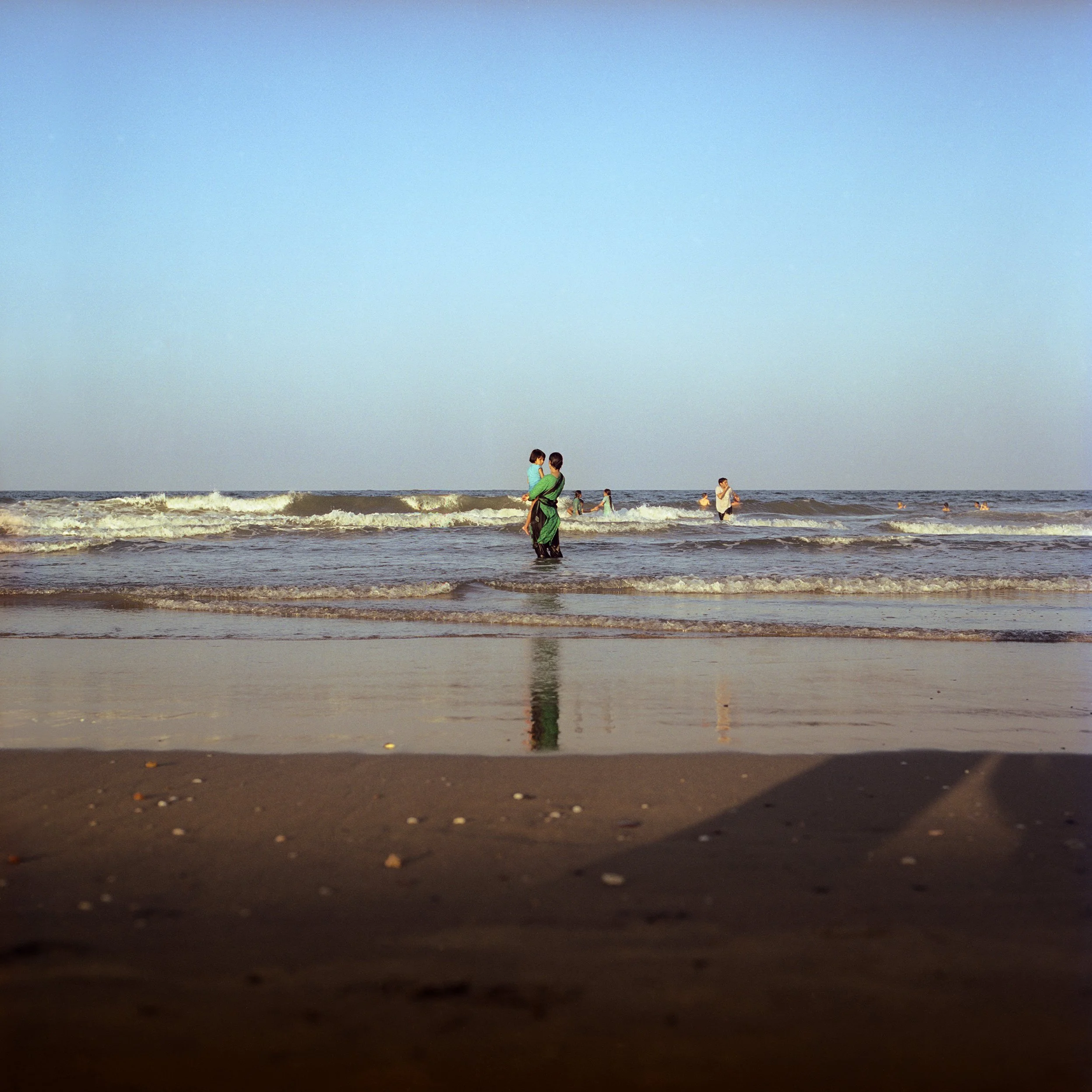 People enjoying a day at the beach with children playing in the water and a parent carrying a child, under a clear blue sky.