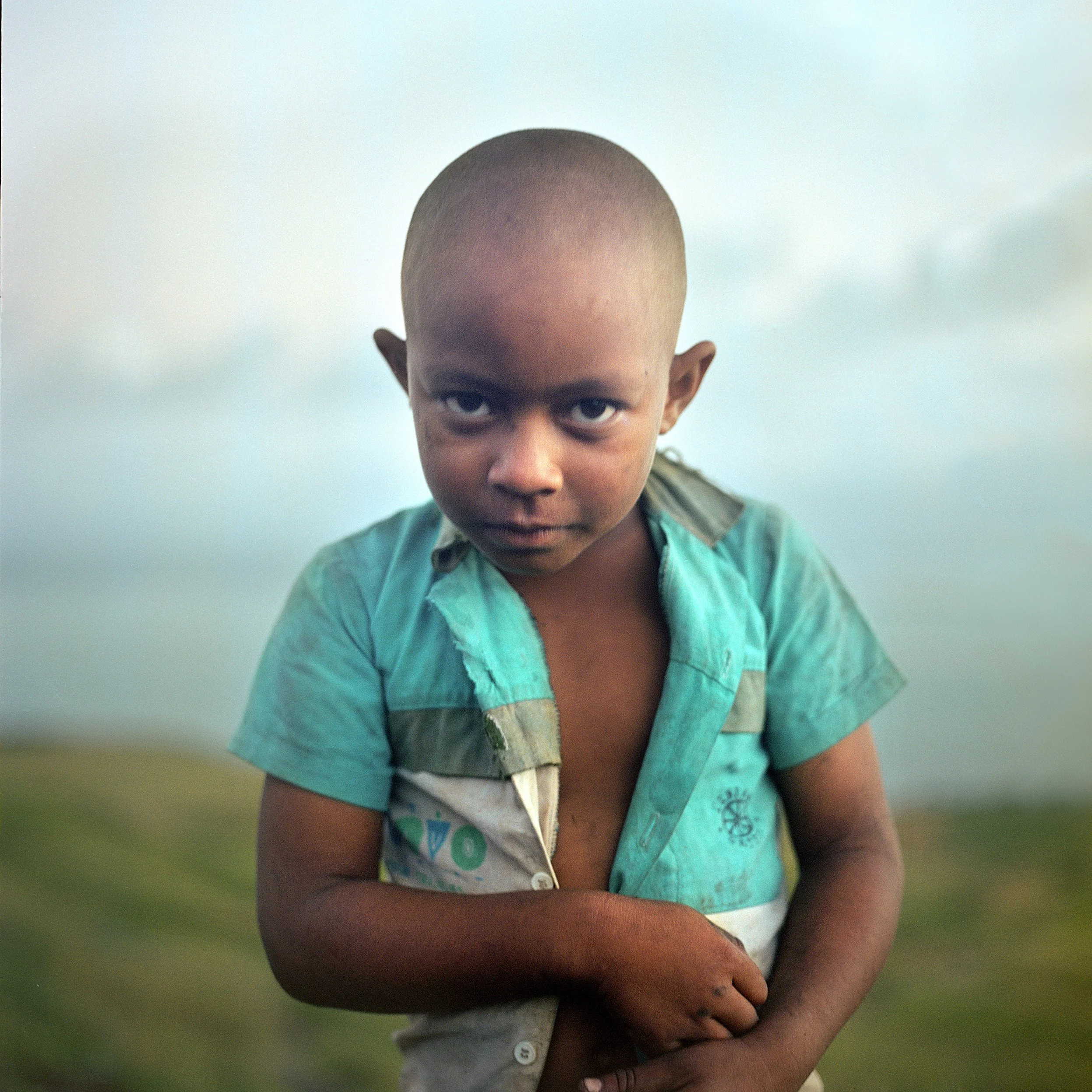 In Rodrigues Island, Mauritius, A boy with a shaved head, wearing a turquoise shirt, standing outdoors against a city sky background.