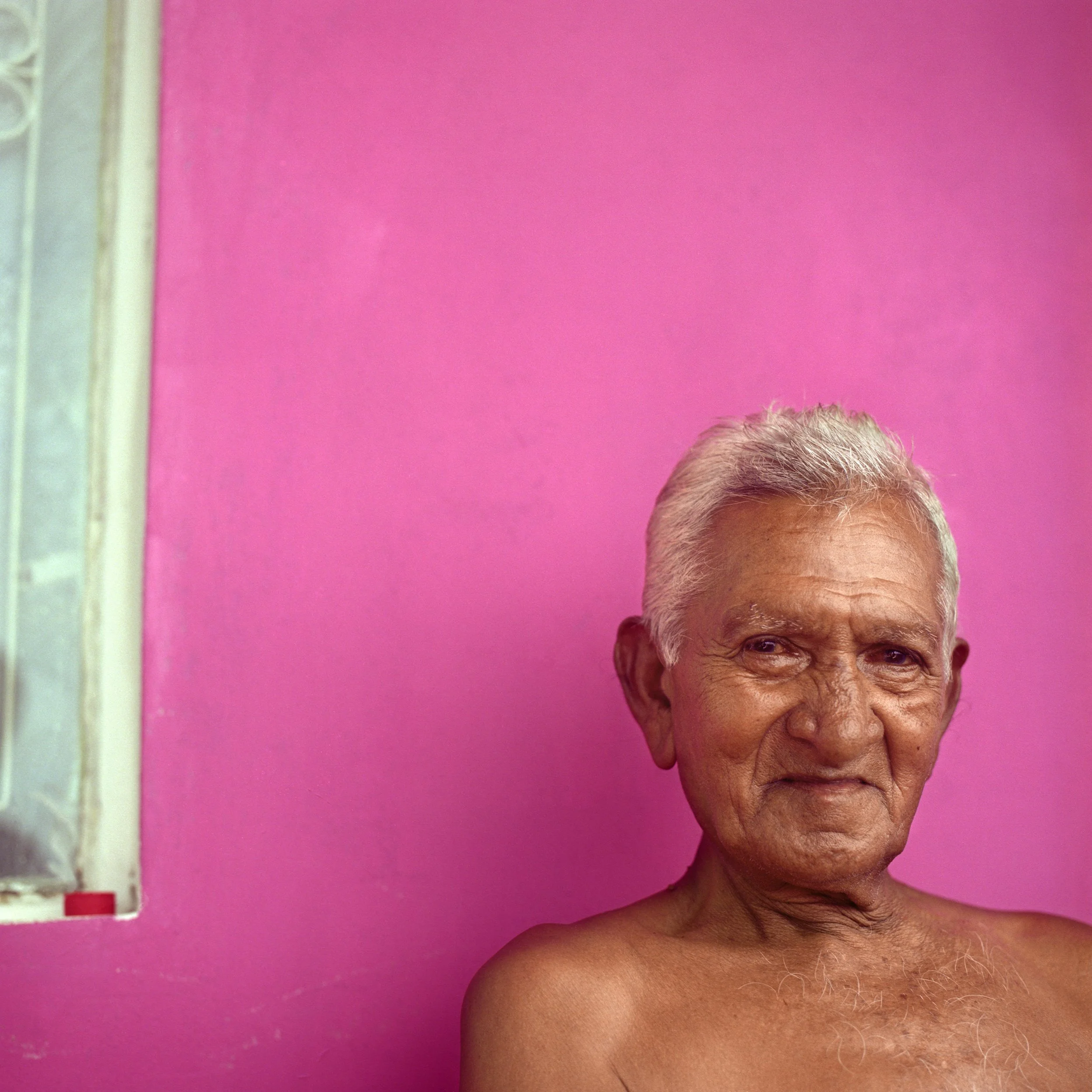 In Mauritius, East Coast, An elderly man with silver hair and a tanned complexion is shirtless, sitting against a pink wall with a window on the left side of the image.