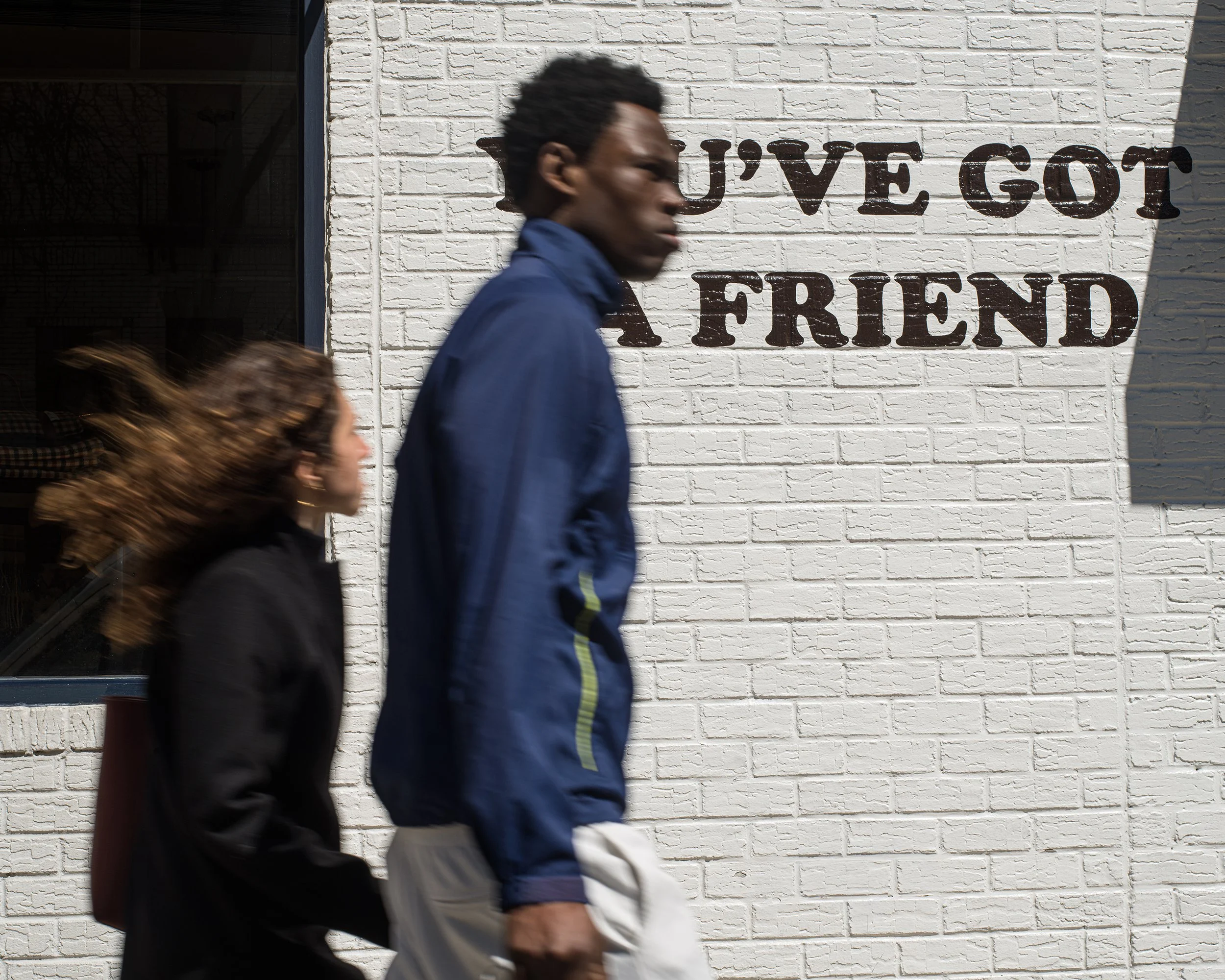 In New York Manhattan, Two people walking past a white brick wall with a sign that reads "I've got a friend".