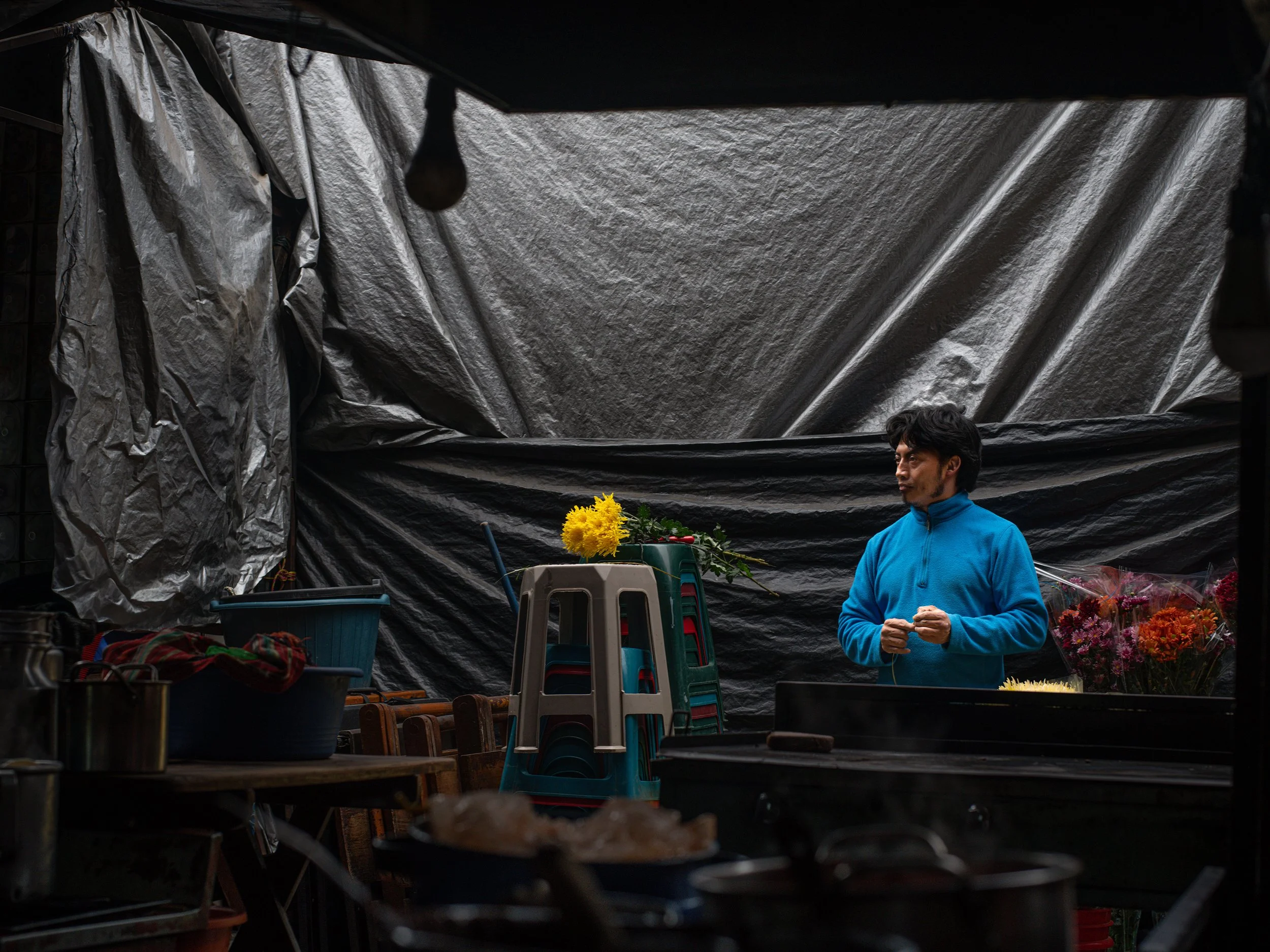 In Chichicastenango market. Guatemala, A man in a bright blue jacket stands in a flower stand with artificial and real flowers, under a tarp-covered structure.