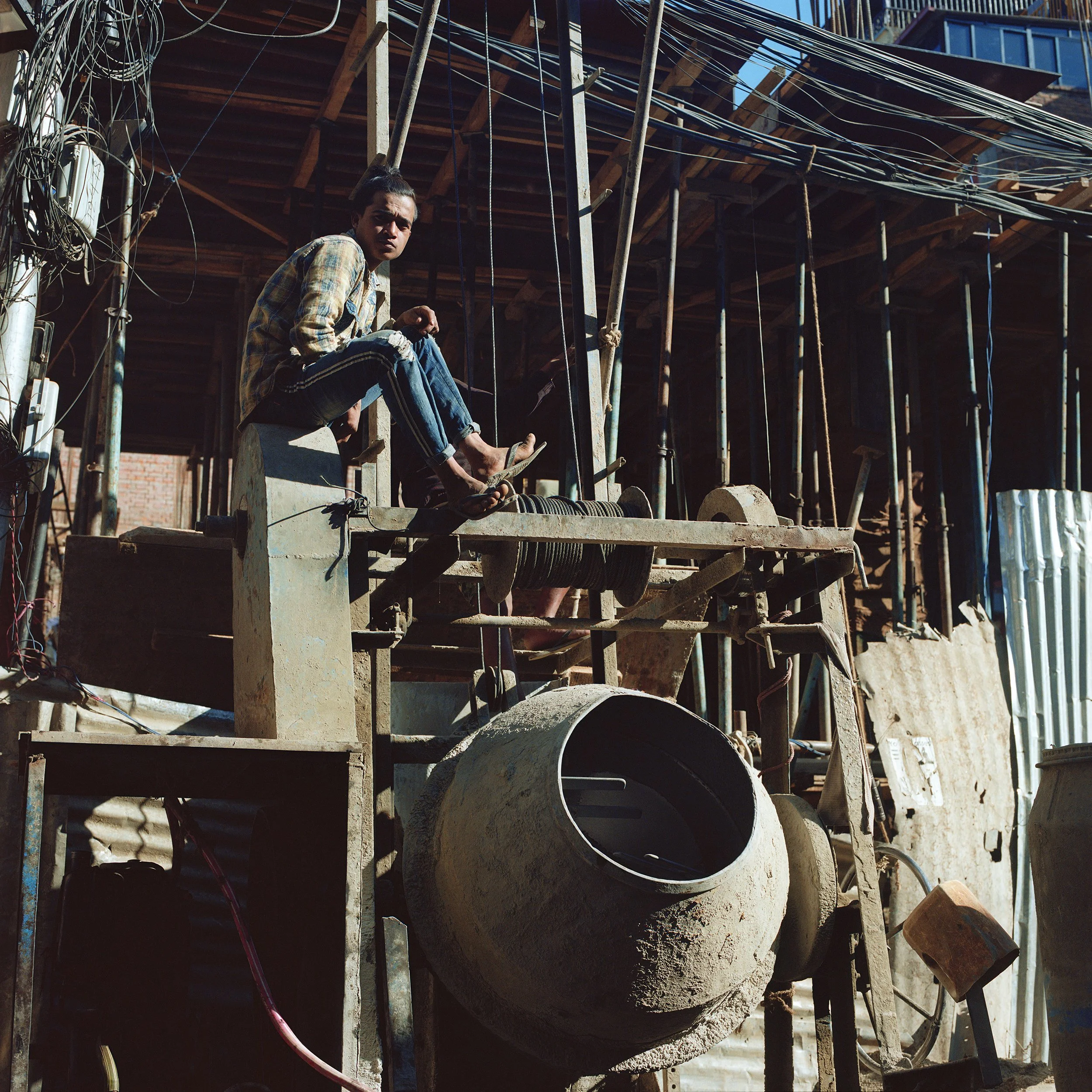 In Katmandu, A young man sitting on a construction site surrounded by building materials and scaffolding, with a large cement mixer in the foreground.