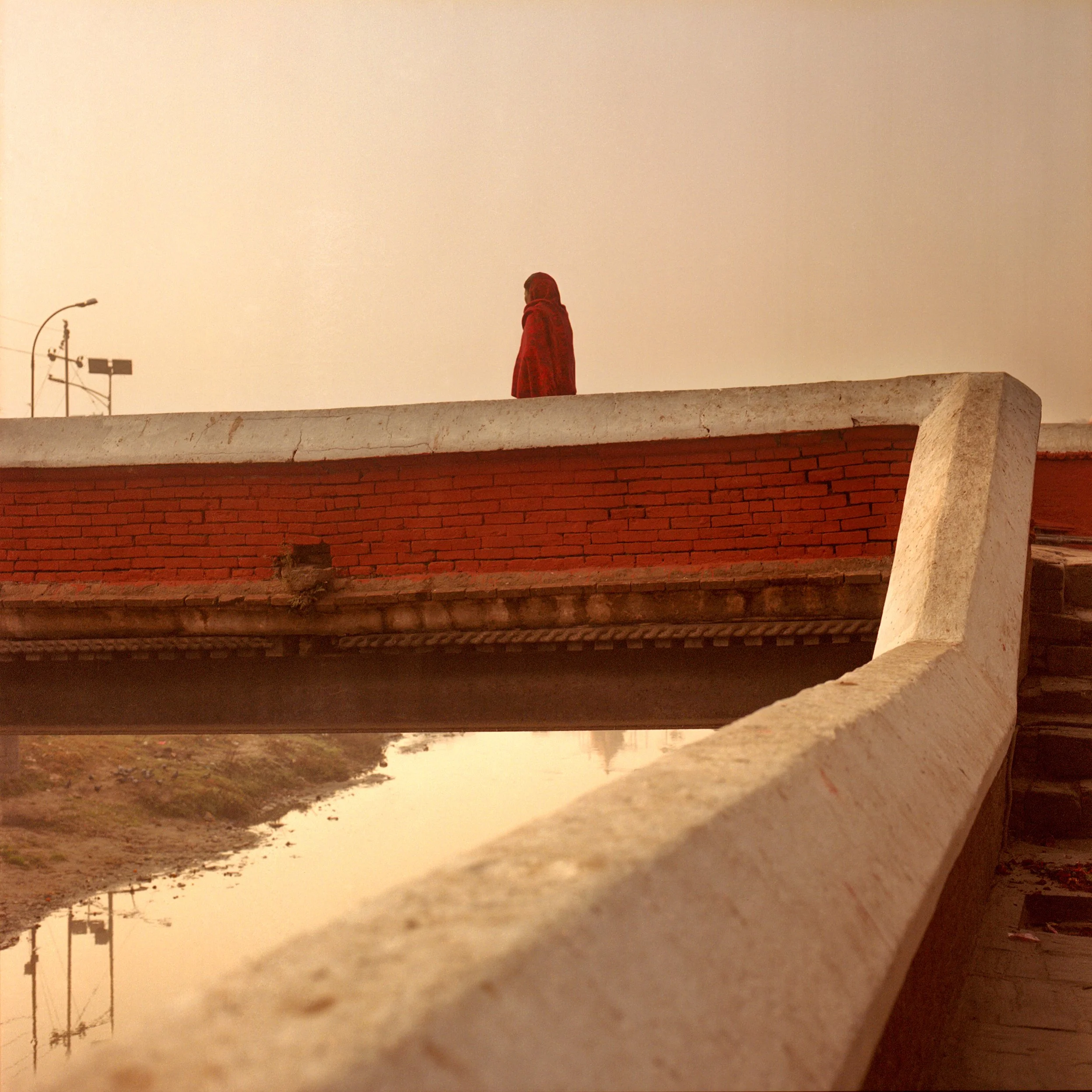 In Kathmandu, A woman in a red coat and headscarf standing on a bridge at sunset, with water and bridge supports below and a clear sky overhead.