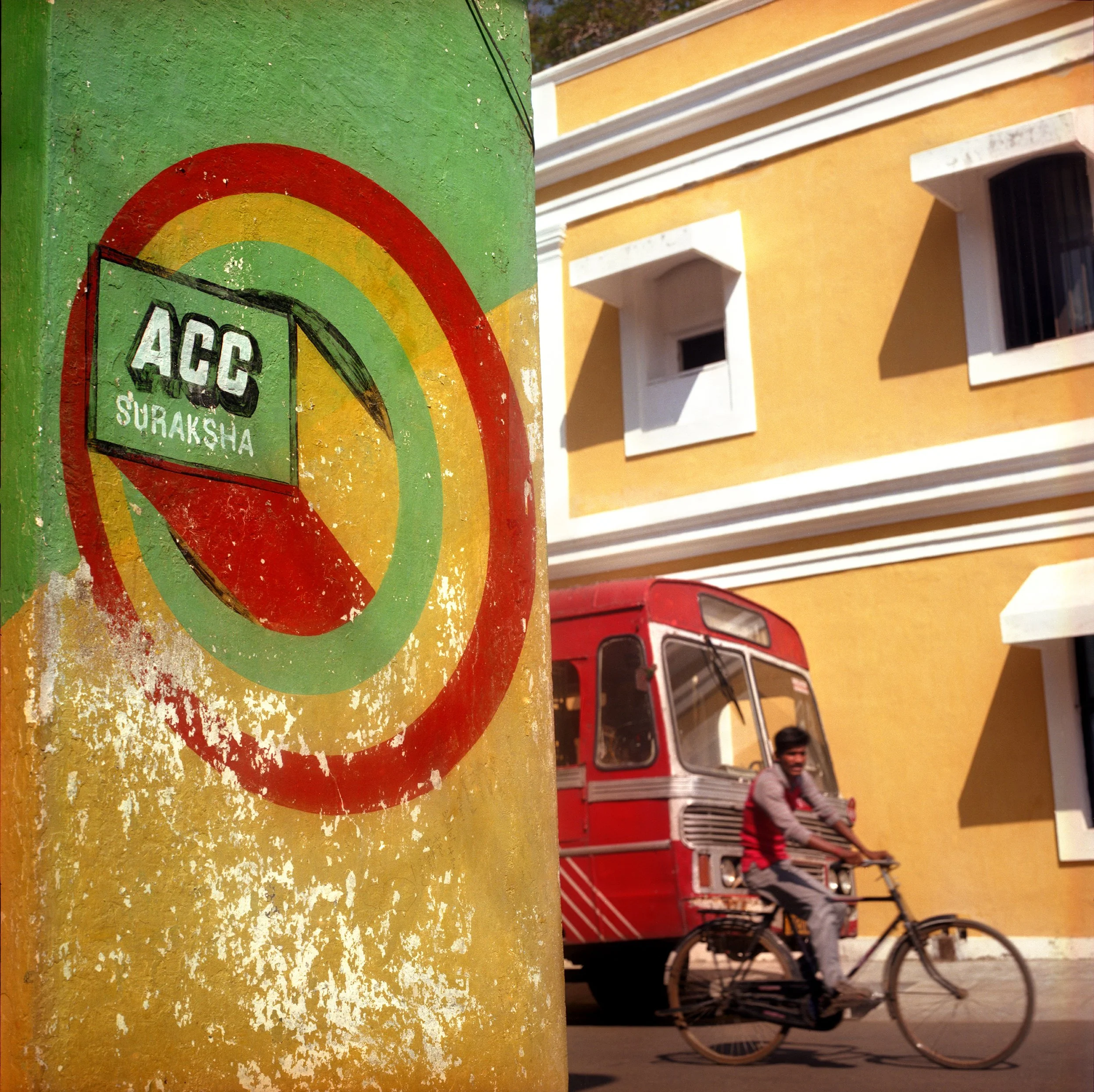 A colorful painted wall with a sign that reads 'ACC SURAKSHA' and a no-smoking symbol. In the background, a man is riding a bicycle past a yellow building with white window frames, and a red auto rickshaw is visible.