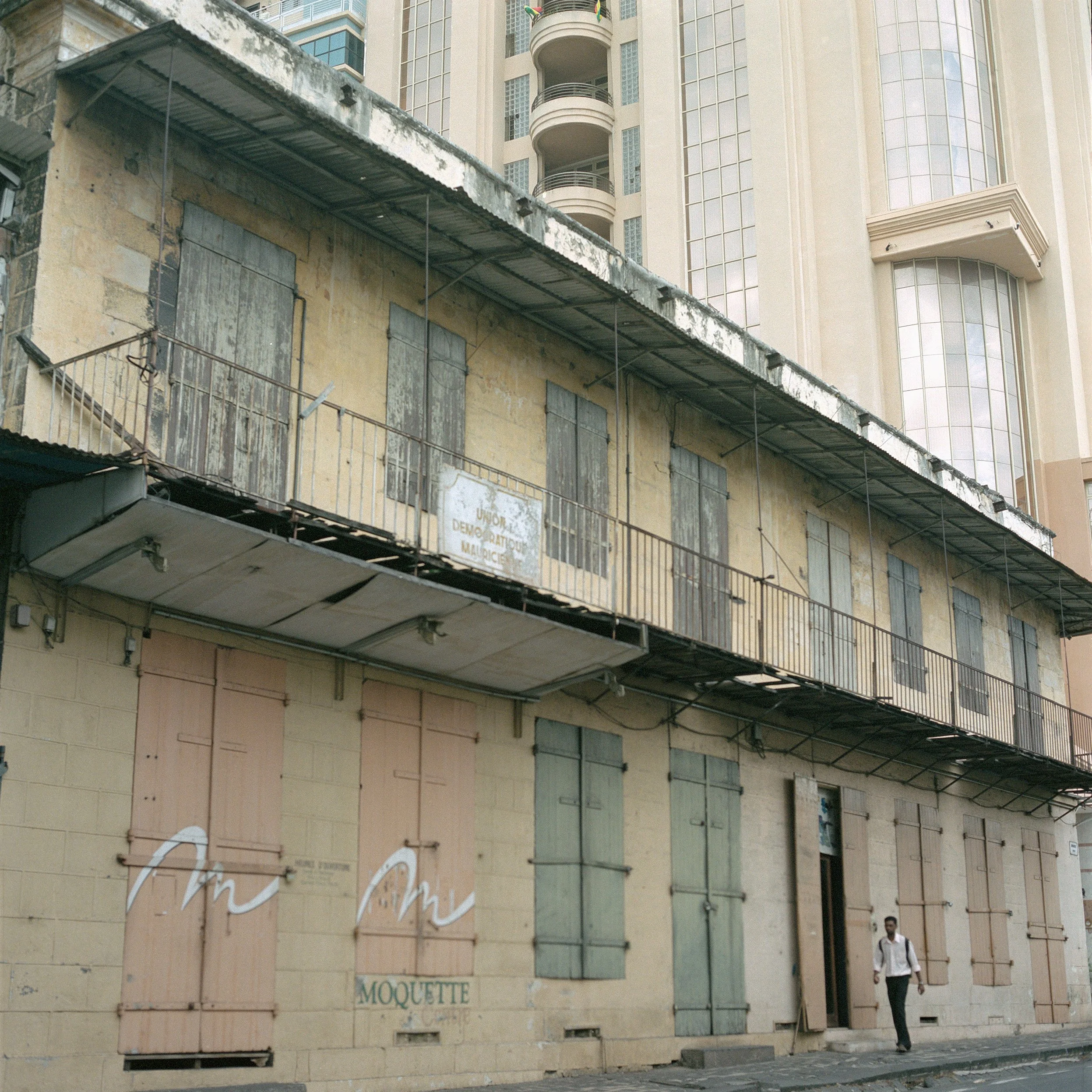 In Mauritius, Port-Louis, An old, abandoned building with boarded-up windows and doors on a street, with a modern high-rise building in the background and a man walking past.