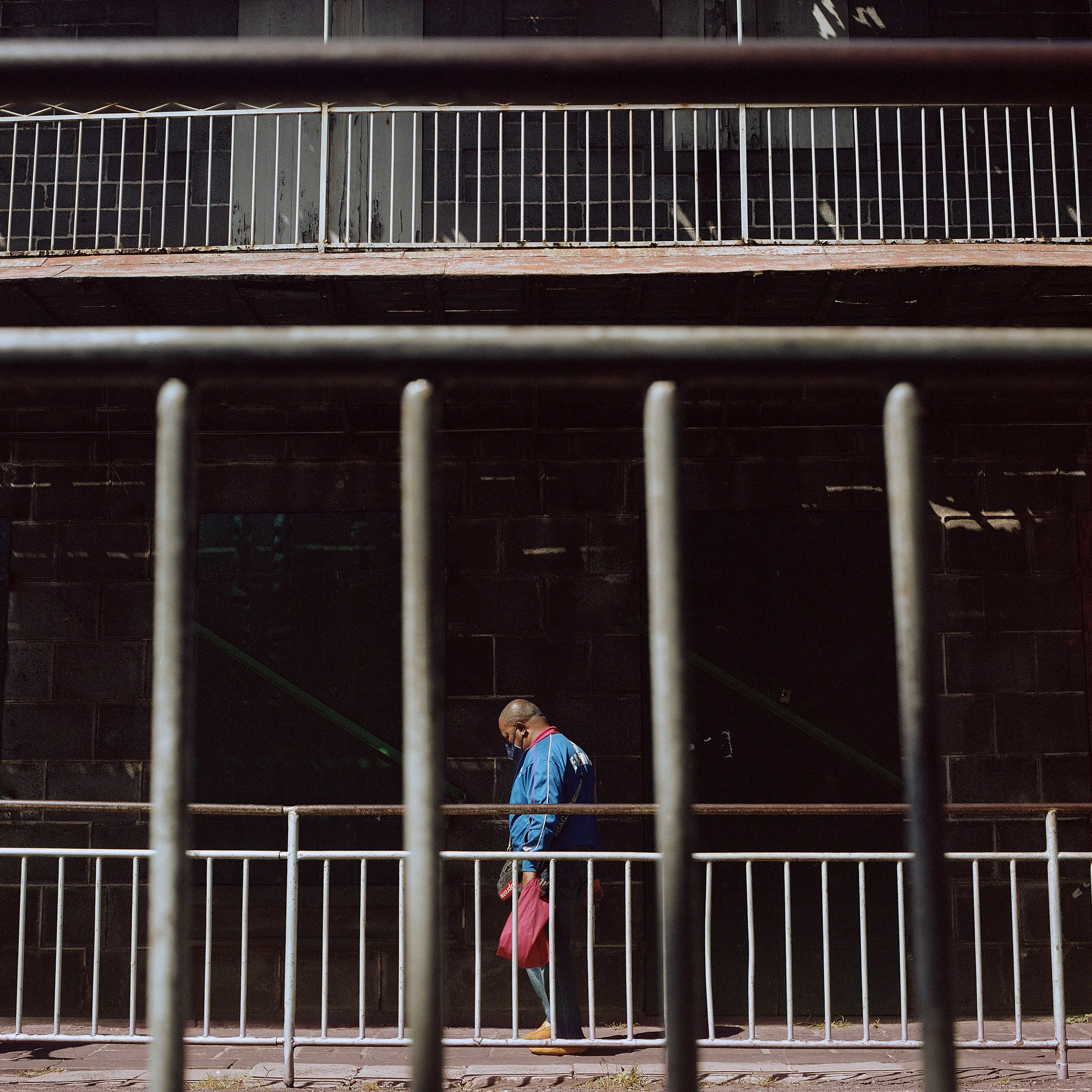 A man wearing a face mask and a blue jacket stands behind a metal railing, holding a pink bag, in front of a dark brick wall.