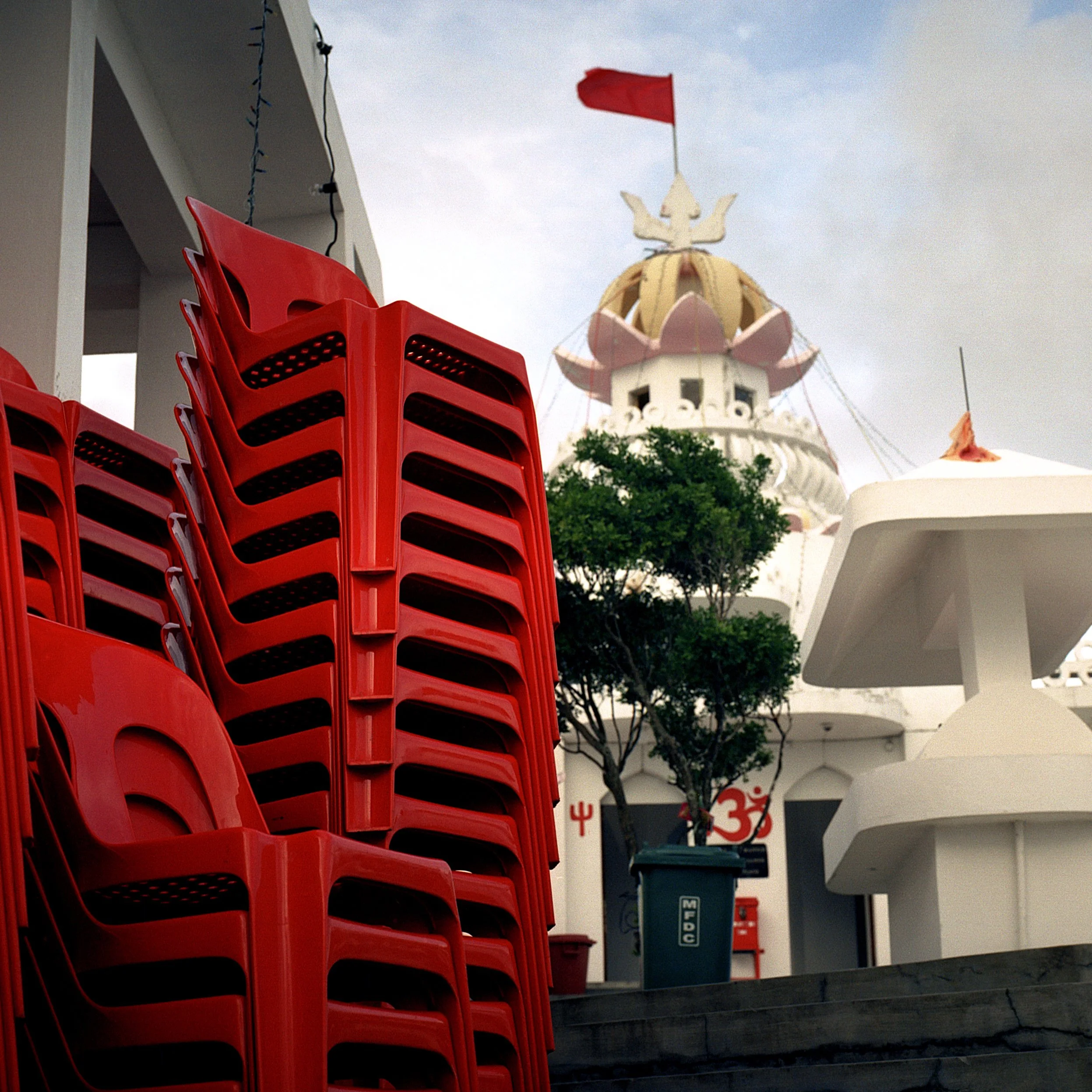In Mauritius, Poste de Flack, A stack of red plastic chairs on a flat surface, with a temple in the background having a golden dome with a red flag on top. The scene appears to be outdoors with a cloudy sky.