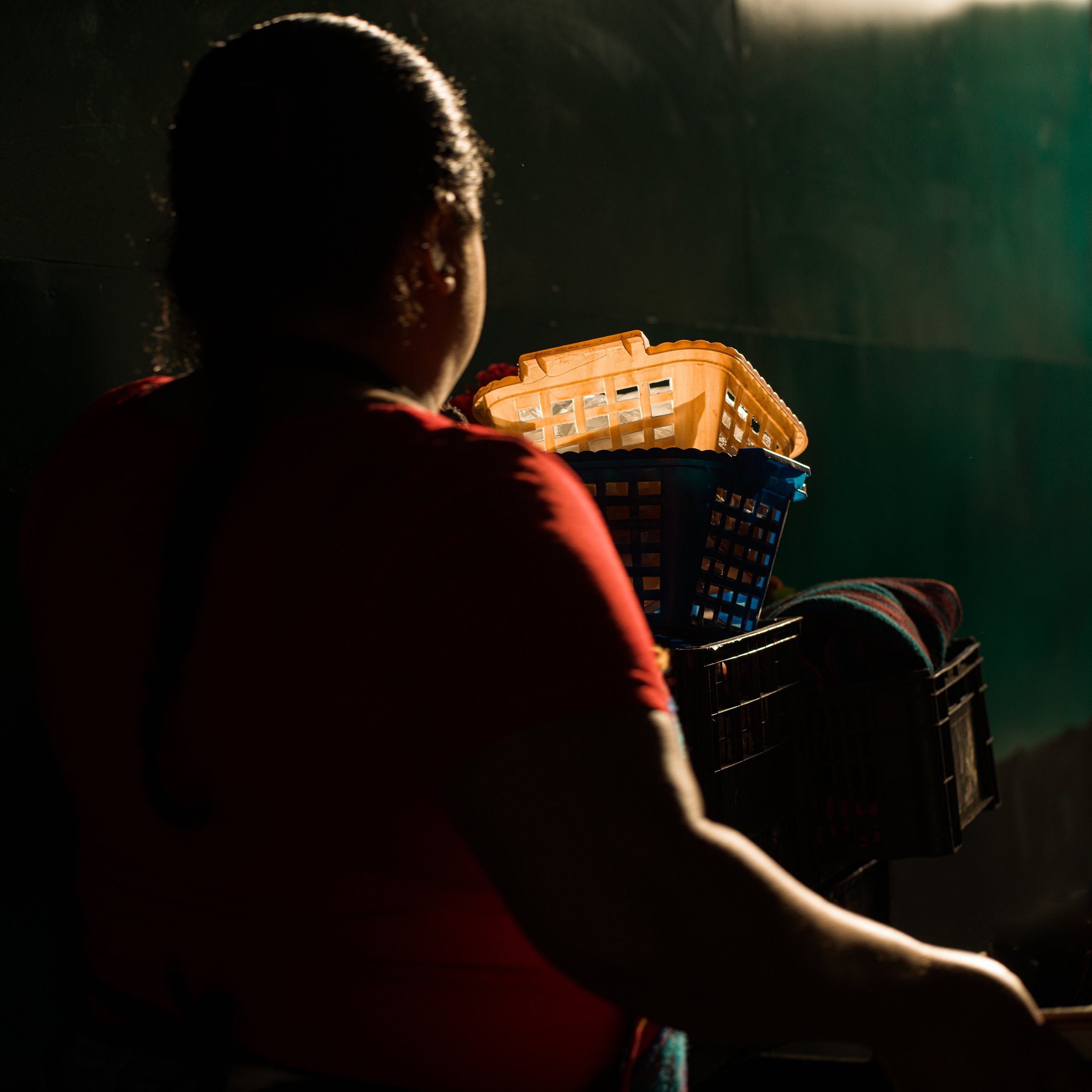 	
In Antigua, Guatemala, market, A woman with braided hair, wearing a red shirt, sitting with her back to the camera in a dimly lit room, holding a basket of laundry, with sunlight casting a glow around her.