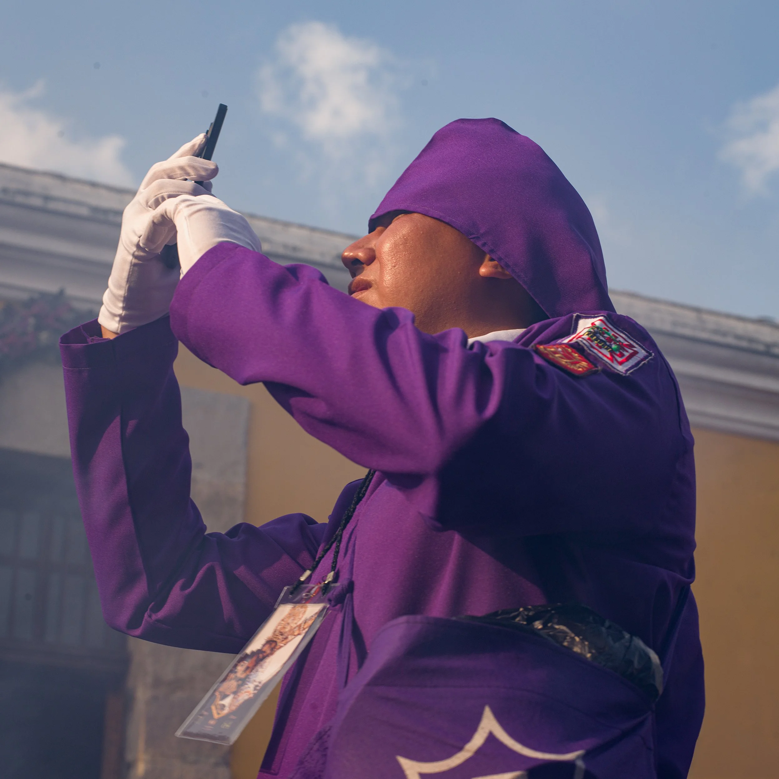 In Antigua, Guatemala, Alfombras, A person dressed in a purple jacket and hood, wearing a white glove, is taking a photograph with a smartphone under a partly cloudy sky.