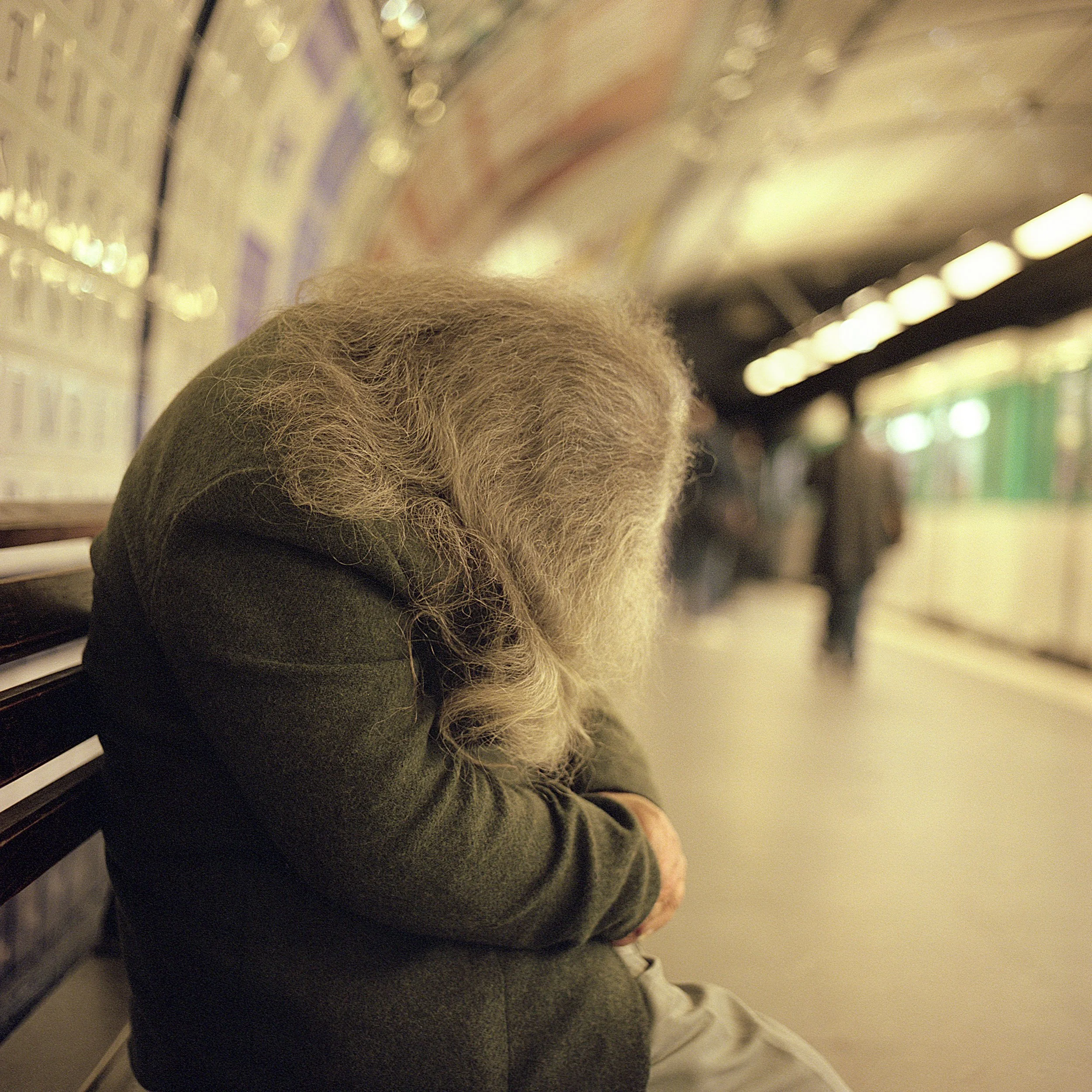 An elderly person in Paris Subway with long gray hair sitting alone on a bench in a subway station, looking down with their head bowed, in a blurry background with other passengers walking.