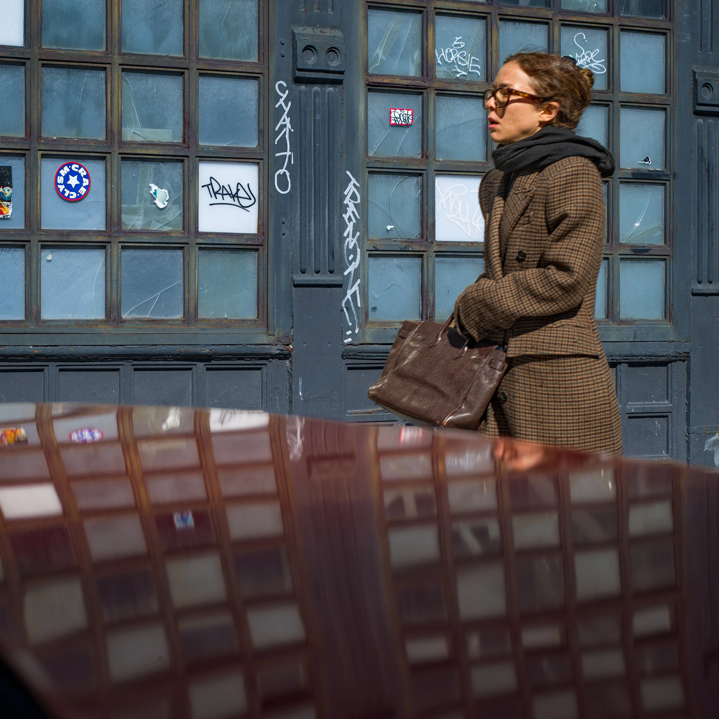 	
In New York Manhattan, A woman in a brown checkered coat and glasses standing outside in front of a graffiti-covered metal garage door, holding a brown leather bag.