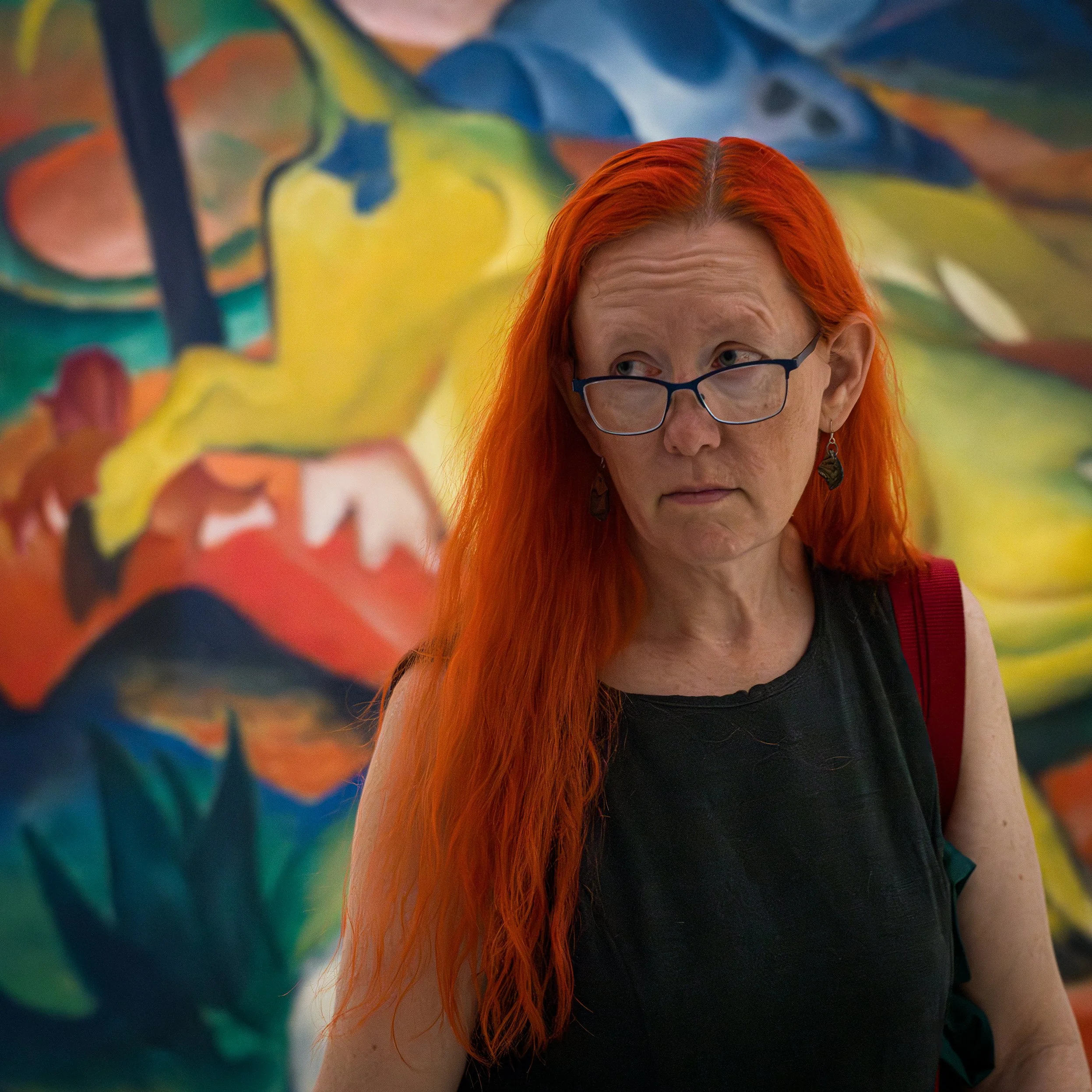 In New York Manhattan, Guggenheim Museum, A woman with long red hair, wearing glasses and earrings, looking to her left in front of a colorful abstract painting.