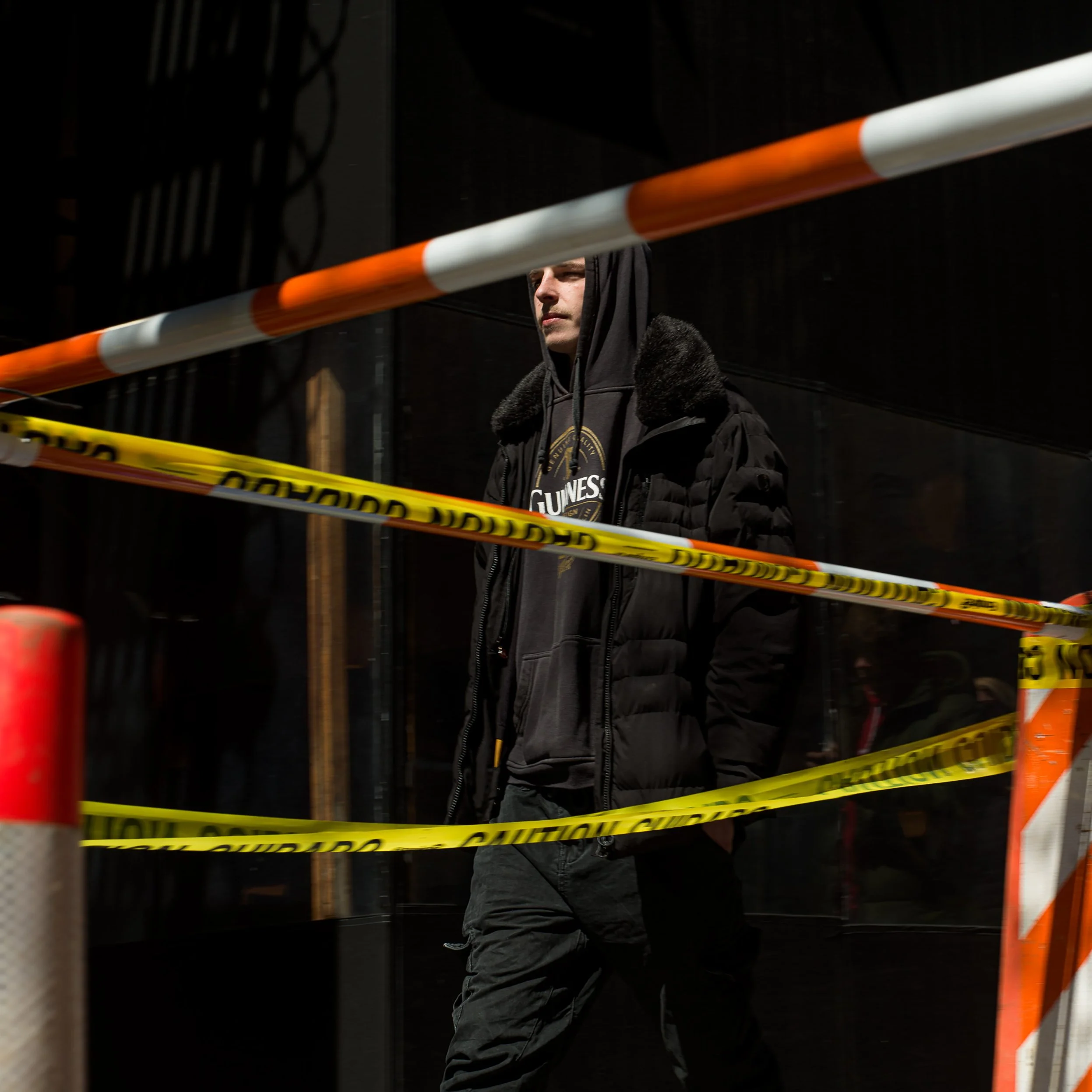 In New York Manhattan, A young man wearing a black hoodie and black jacket walking past construction or restricted area with yellow caution tape.