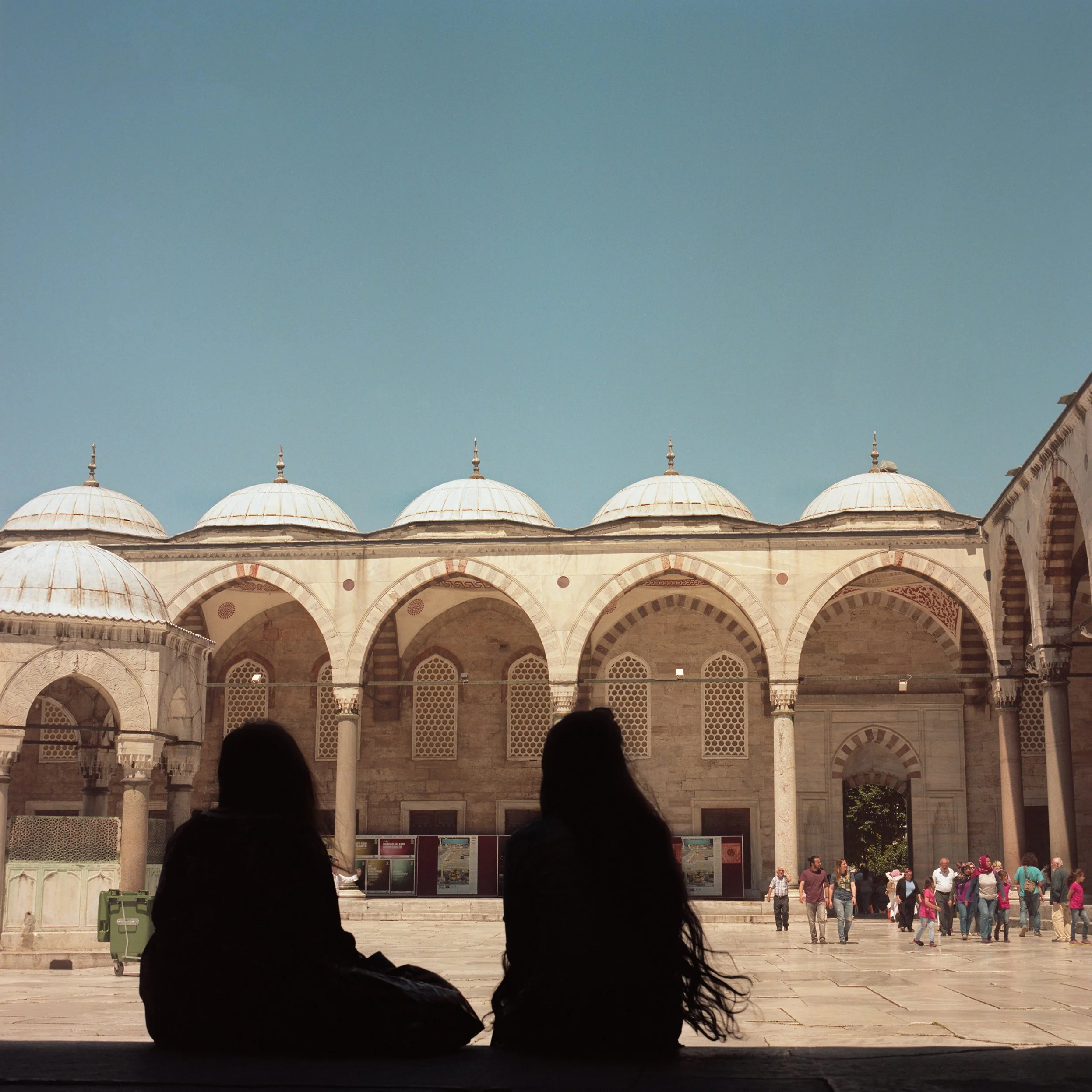In Istanbul, Turkey, People sitting and standing in a courtyard with historic architecture, arches, and domed rooftops under a clear blue sky.