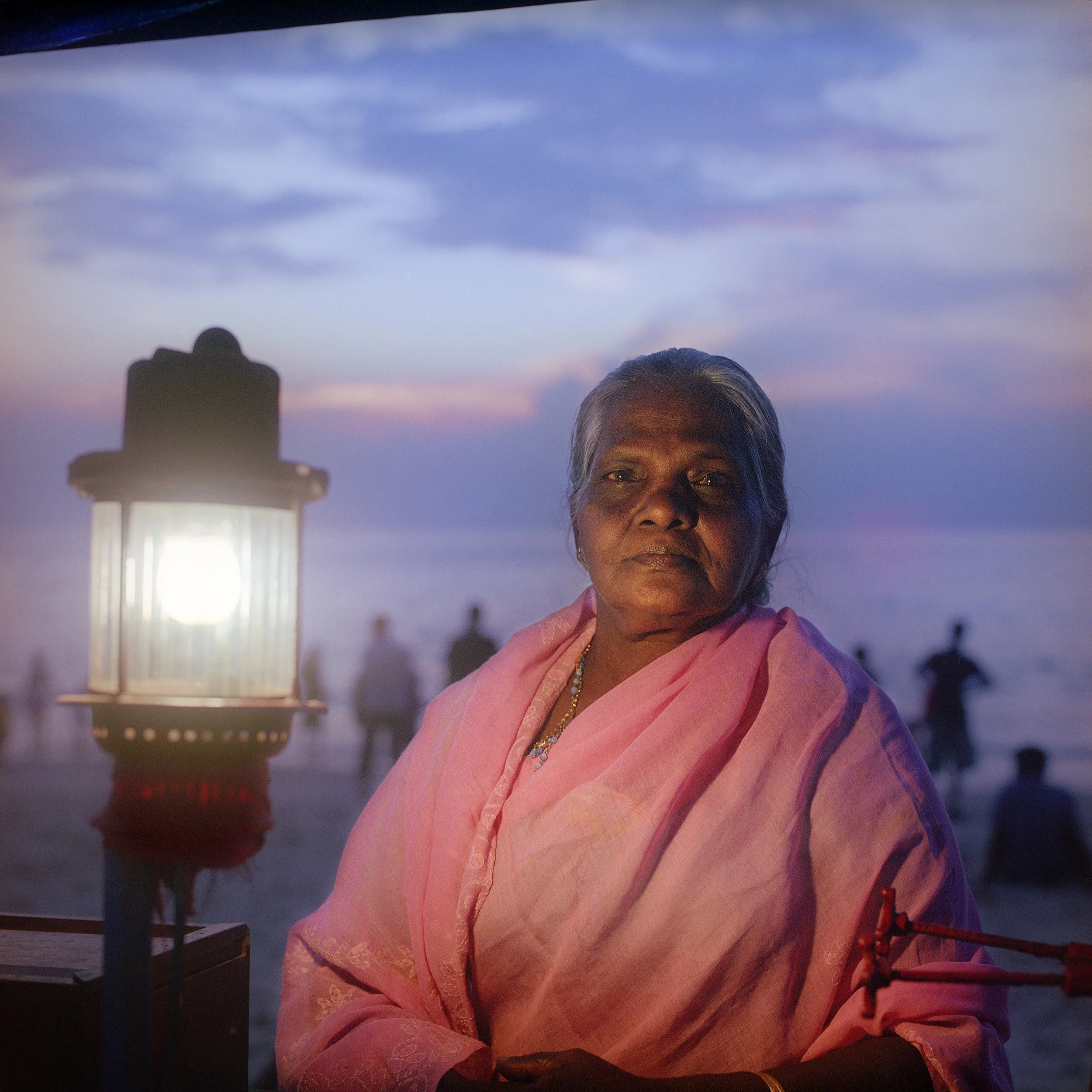 In Kerala, India, Close-up of an elderly woman in a pink traditional outfit, standing outdoors during dusk with a lighthouse in the background and a twilight sky with clouds.