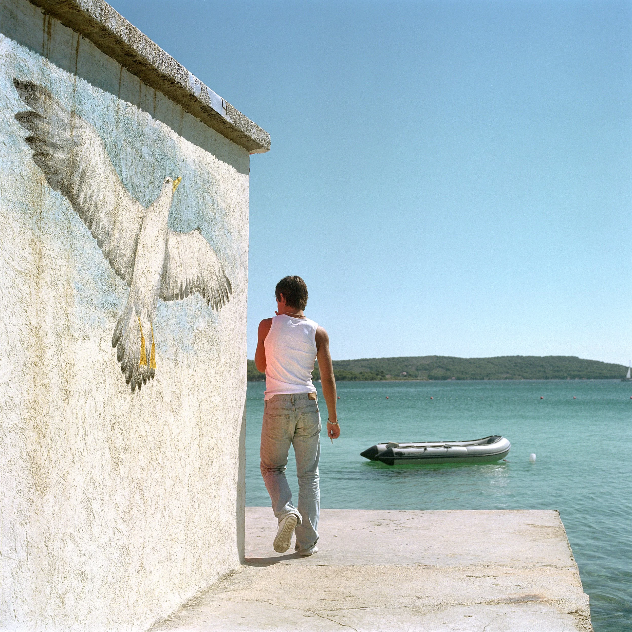 A person walking along a concrete dock next to a white wall with a painted seagull, overlooking a body of water with a small boat floating nearby, under a clear blue sky.