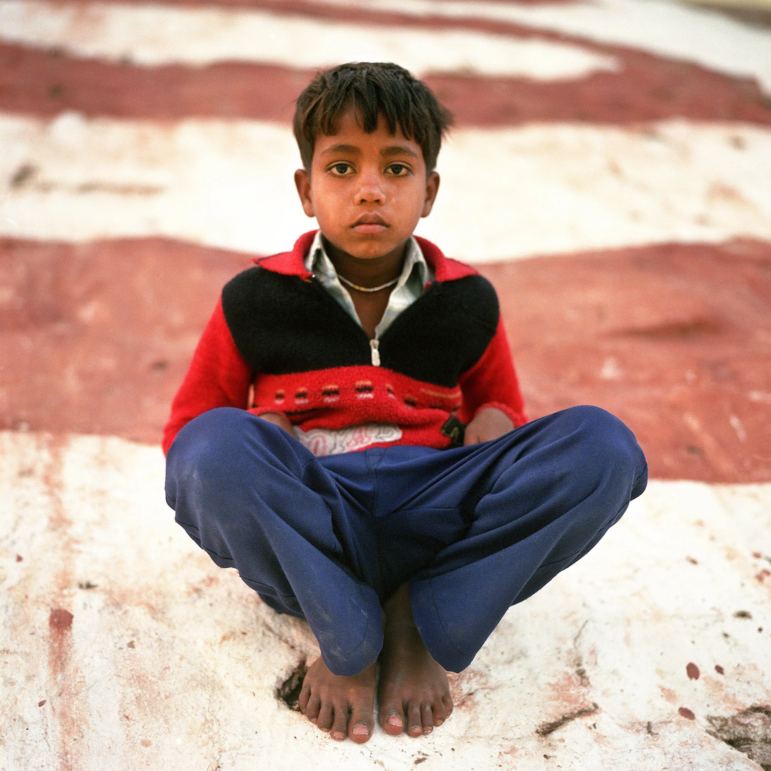 Young boy sitting cross-legged on a stepped surface, wearing a red and black sweater and blue pants, looking directly at the camera.
