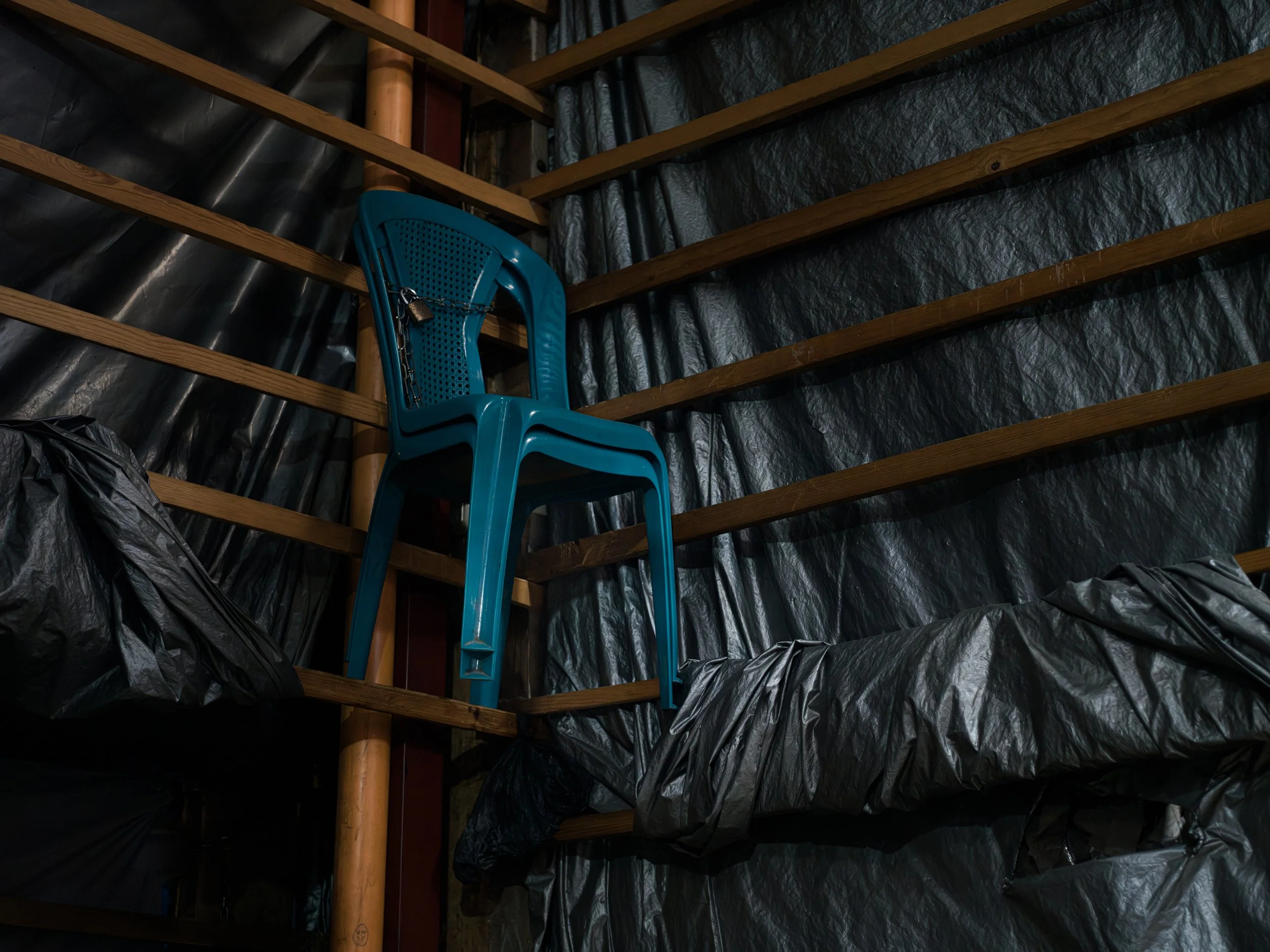 In Chichicastenango market. Guatemala, A blue plastic chair chained to wooden shelves in an unfinished or storage room with black plastic sheeting on the walls.