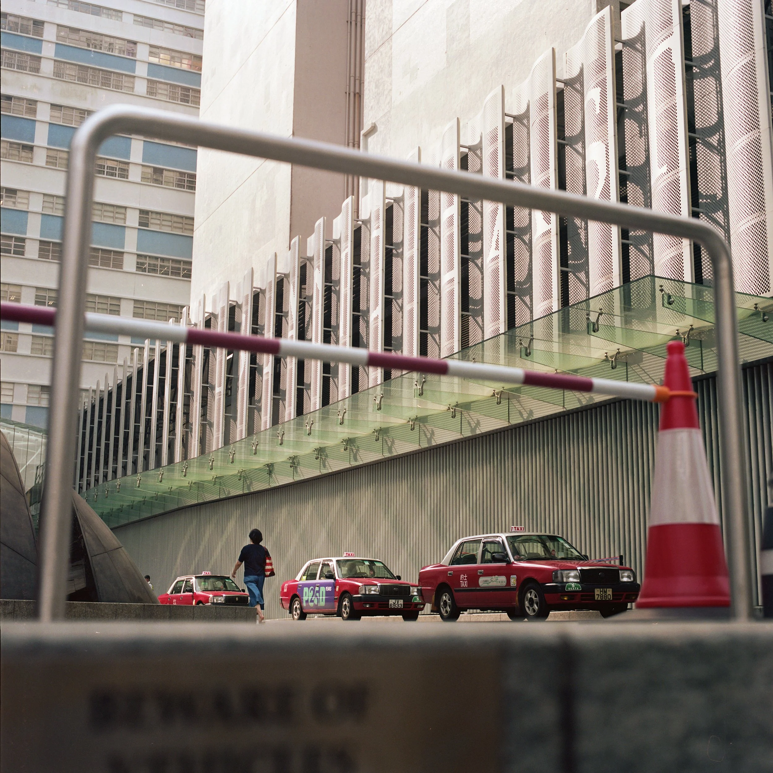 Street scene with red taxis, a person walking, and a modern building with a unique architectural façade in the background. The photo is taken through a parking barrier and traffic cone.