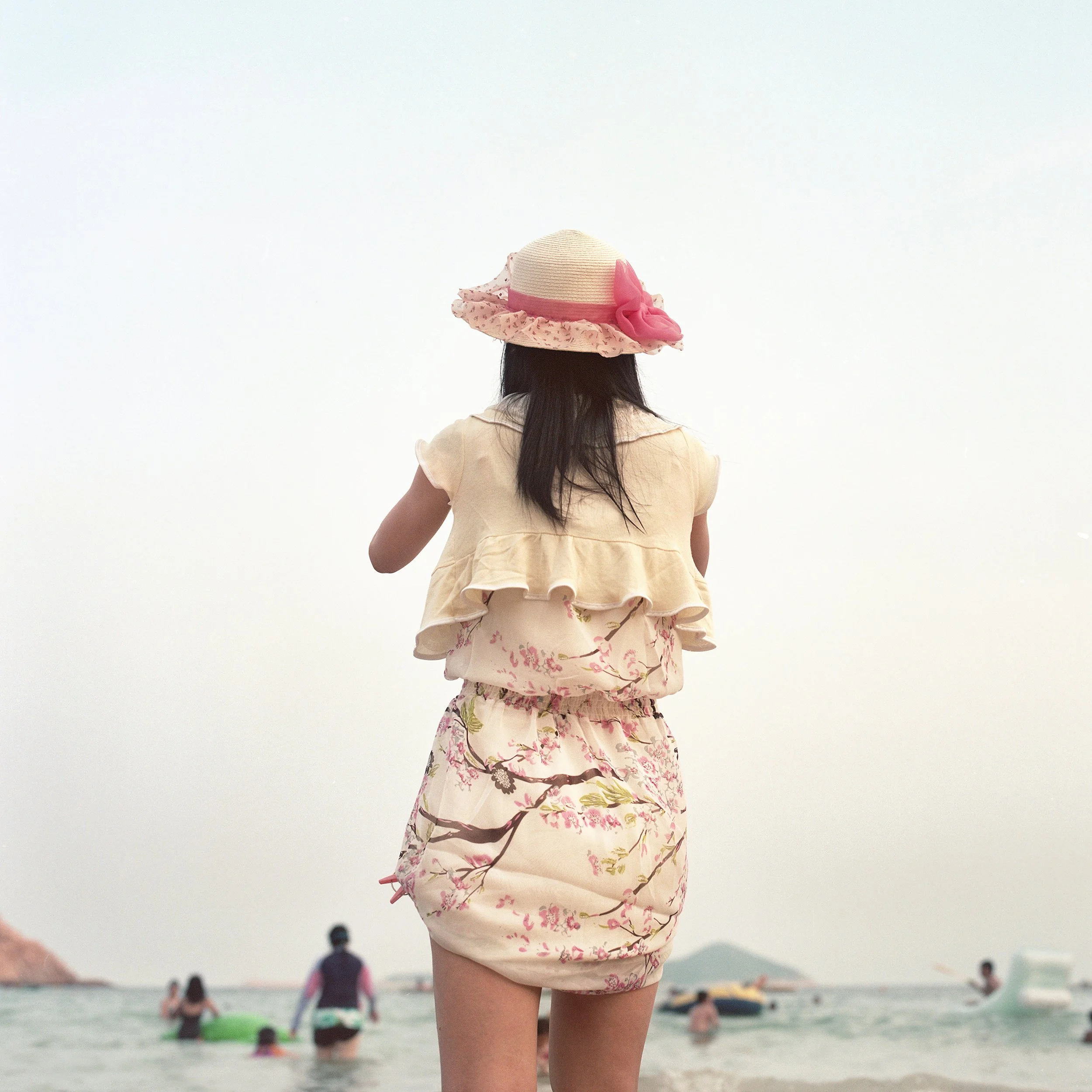 A girl wearing a floral skirt and a beige top, with a wide-brimmed straw hat with pink ribbon, standing on a beach with people swimming and boats in the background.