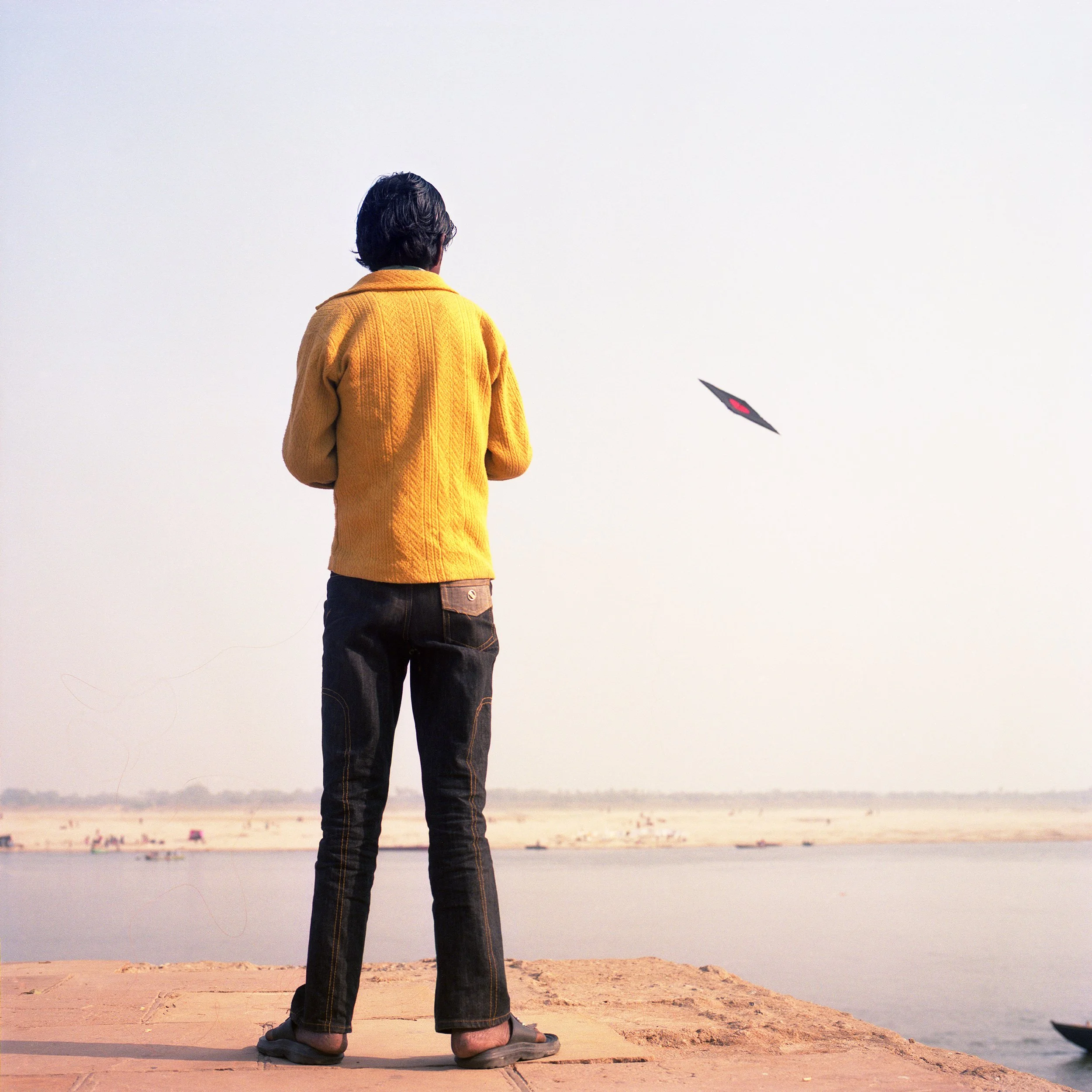 Man standing on riverside watching a kite flying in the sky.