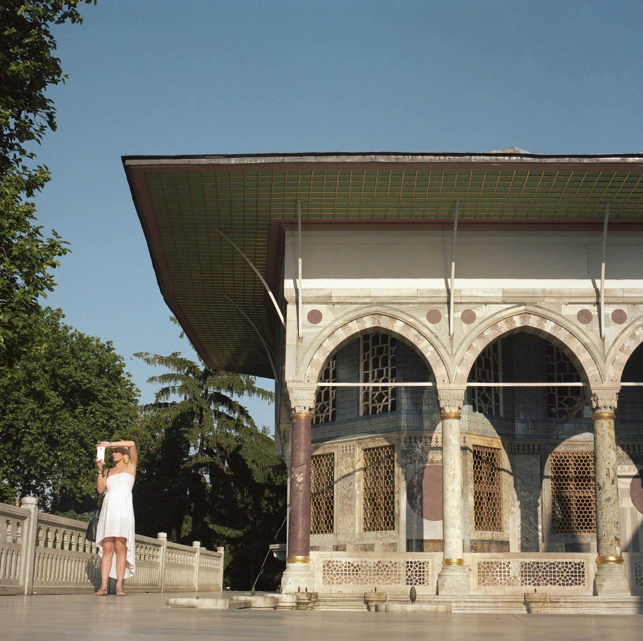 In Istanbul, Turkey, A woman in a white dress taking a photo with a pink phone outside a historical building with arches and columns.