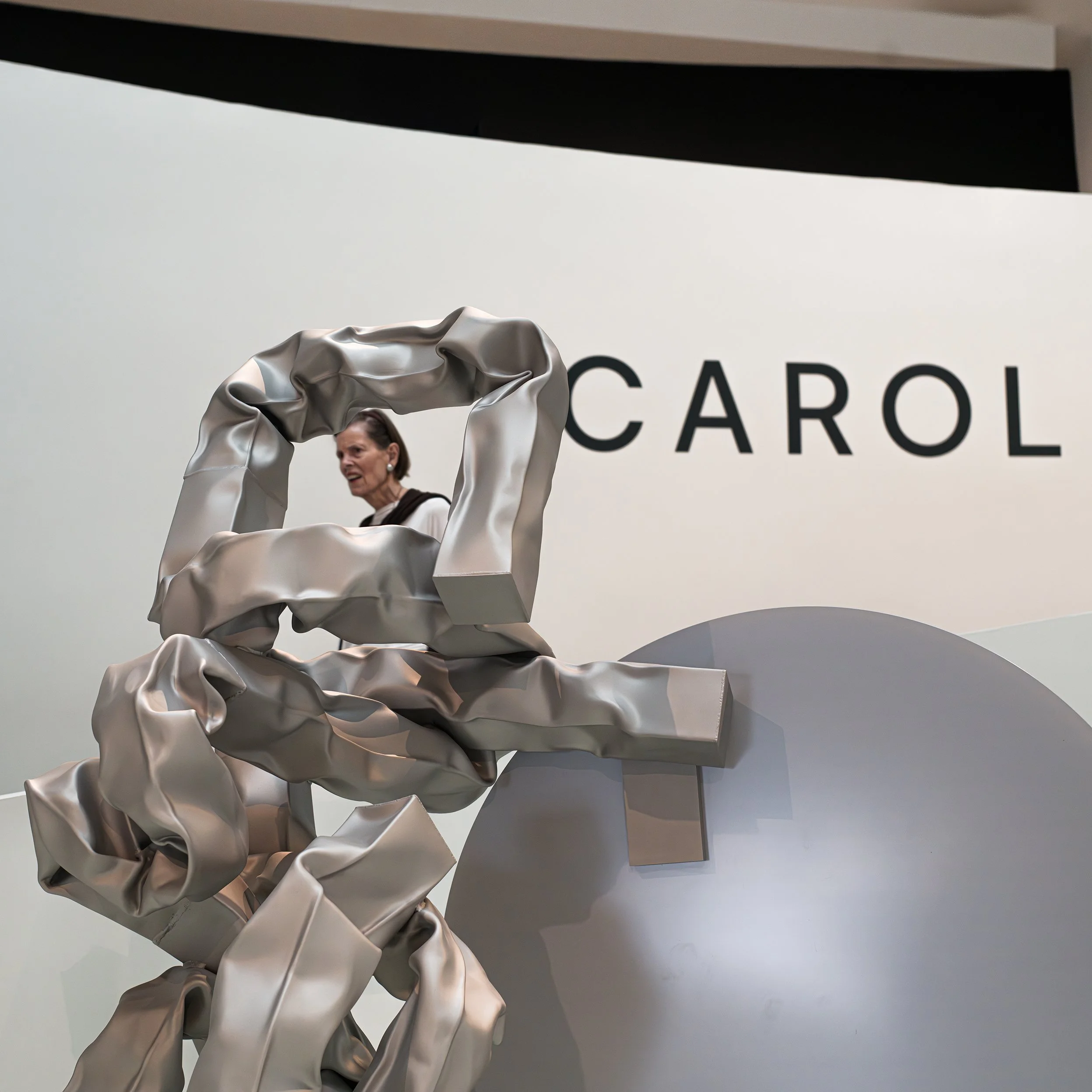 	
In New York Manhattan, Guggenheim Museum, A woman walking past a modern art sculpture resembling a large, crumpled metallic ribbon, with the word 'CAROL' displayed on a white wall in the background.