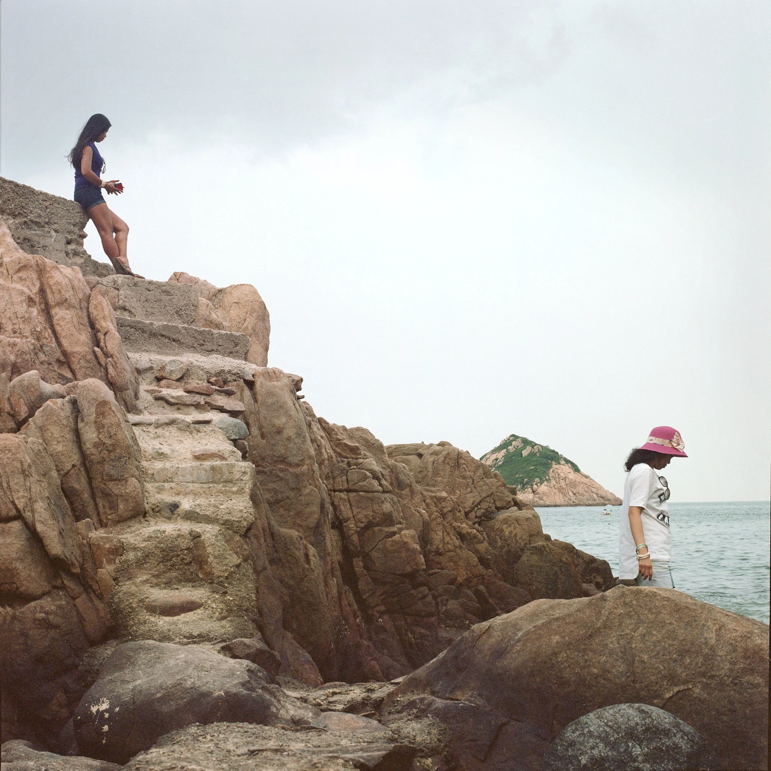 Two women on rocky coastline with ocean in background, one standing on rocks higher up, the other walking along the shore wearing a pink sun hat.